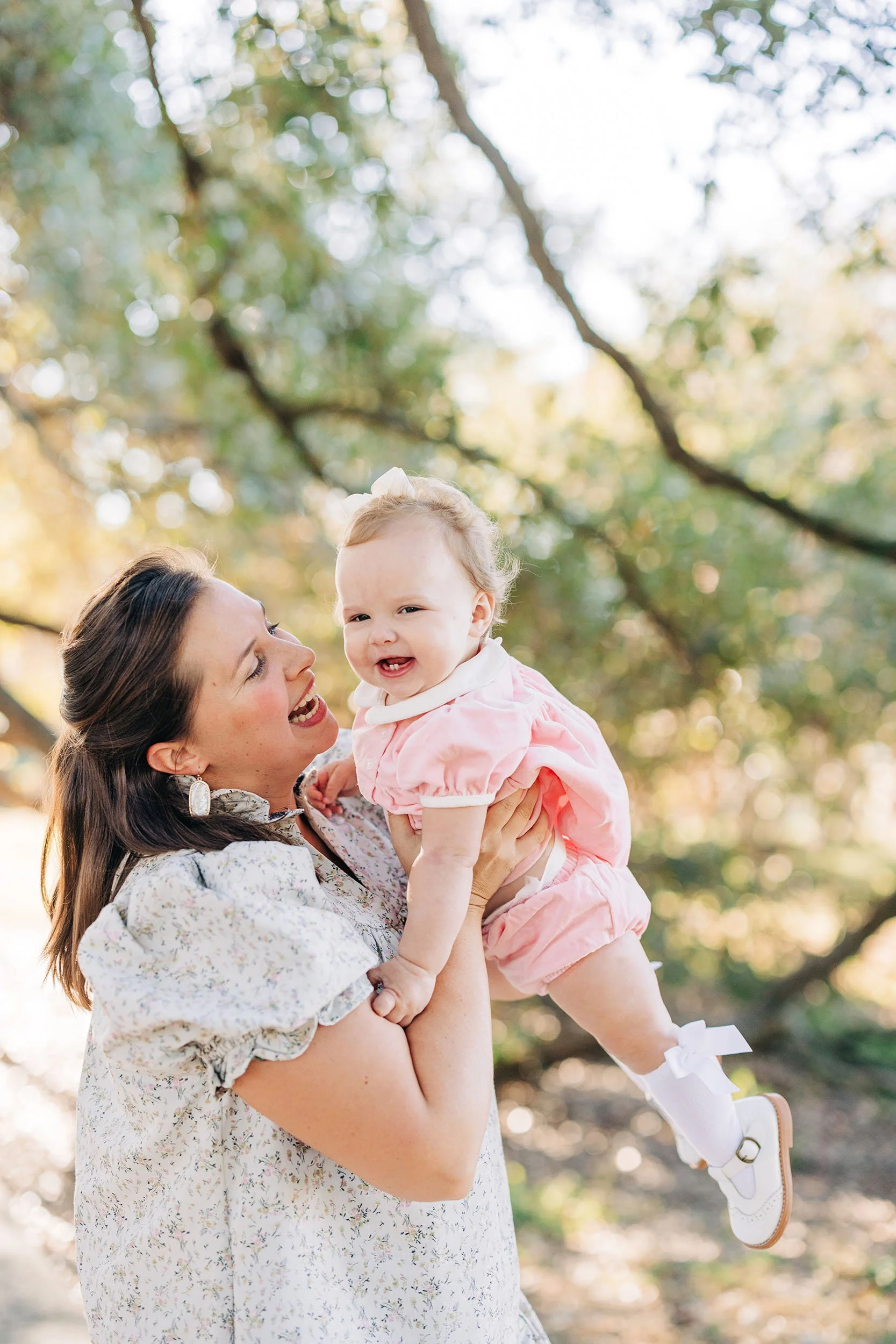 A woman with brown hair, wearing earrings and a gray floral dress, is holding up a smiling baby girl in a pink outfit with white shoes and socks, outdoors with trees and sunlight in the background.