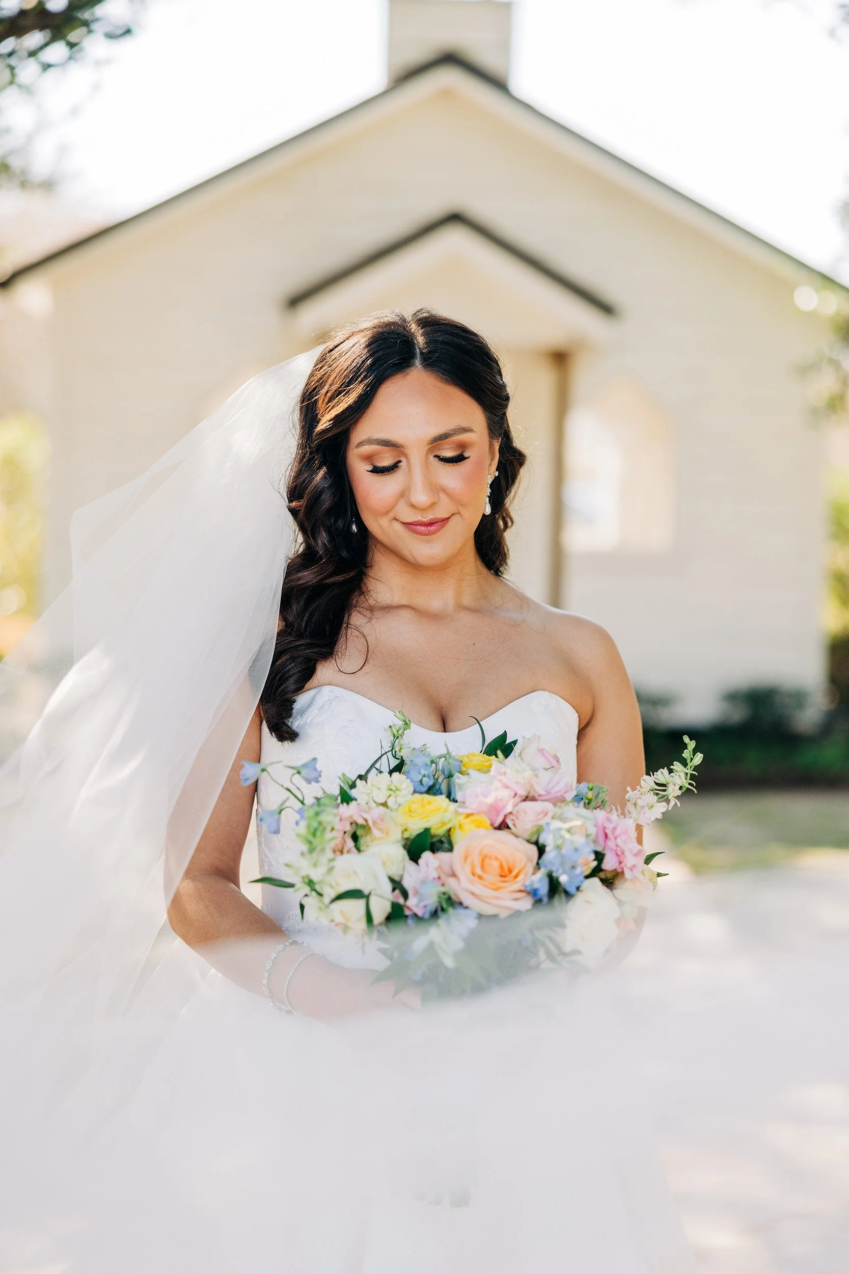 A bride with dark wavy hair wearing a white strapless wedding dress and veil, holding a colorful bouquet, standing outdoors with a yellow house in the background.