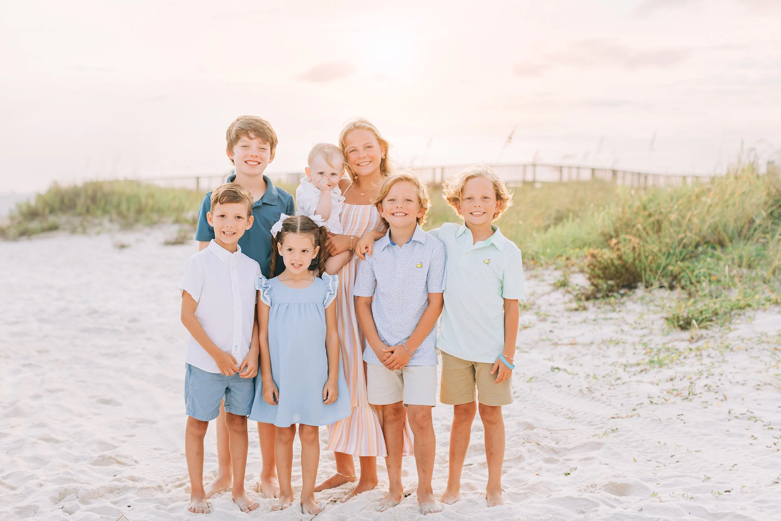 A group of children and a woman standing on a sandy beach with grass and a pier in the background, during sunset or sunrise.