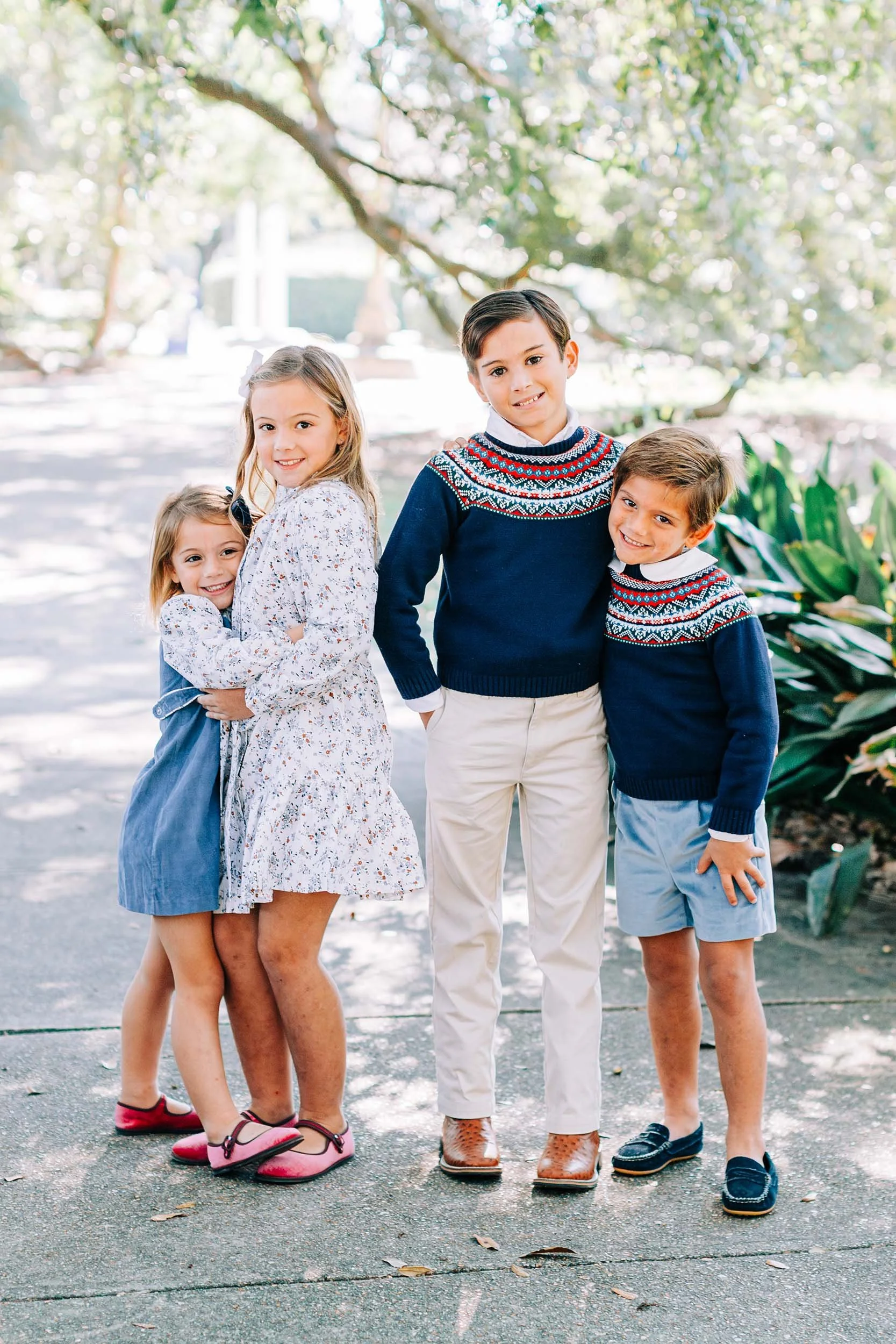 Four children with light skin standing outdoors on a paved path surrounded by trees and greenery, smiling at the camera. The children are dressed in casual clothes, with two boys and two girls, some wearing patterned sweaters and others in dresses or