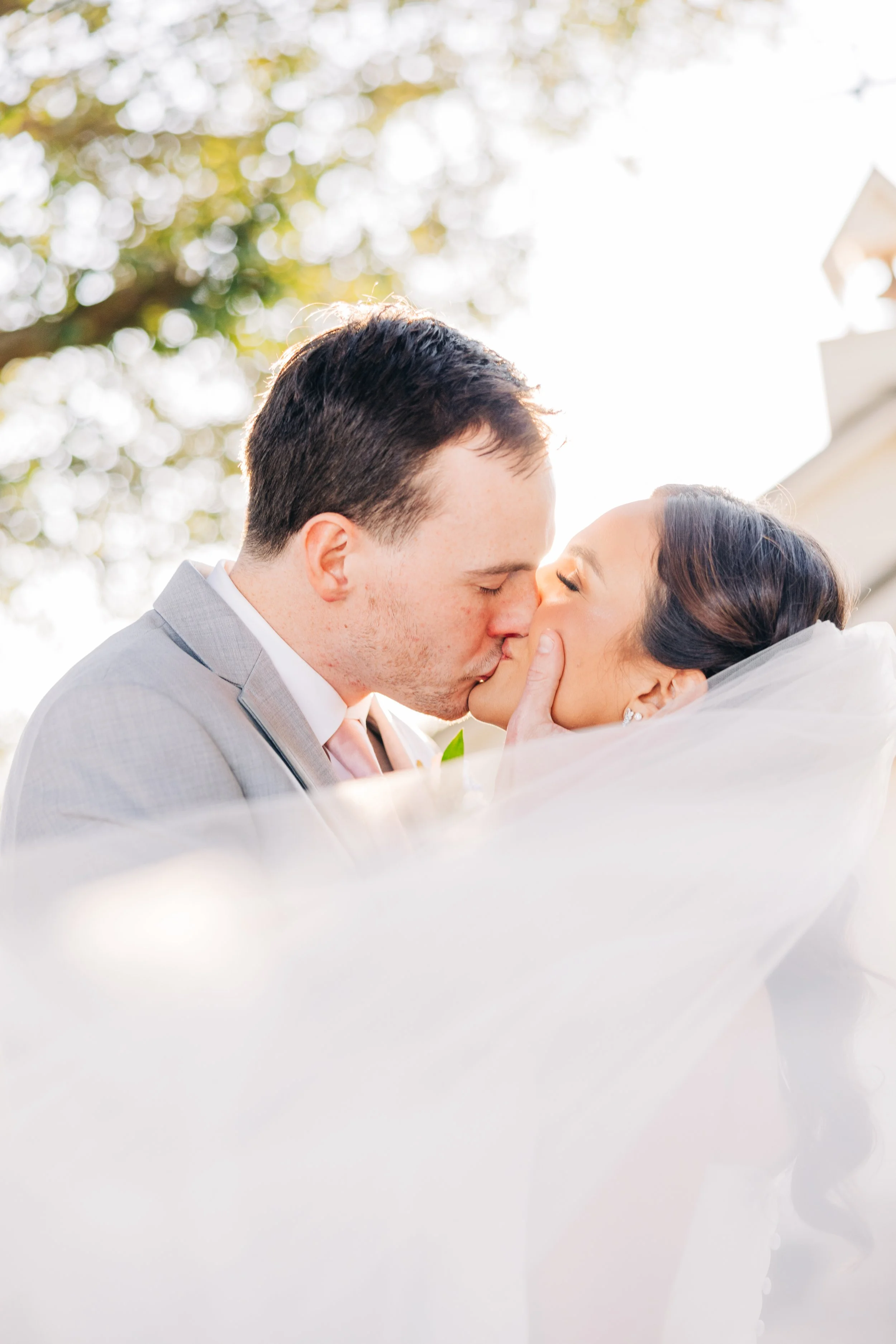 A man and a woman, likely newlyweds, are sharing a kiss outdoors during daylight, with trees and bright sunlight in the background.