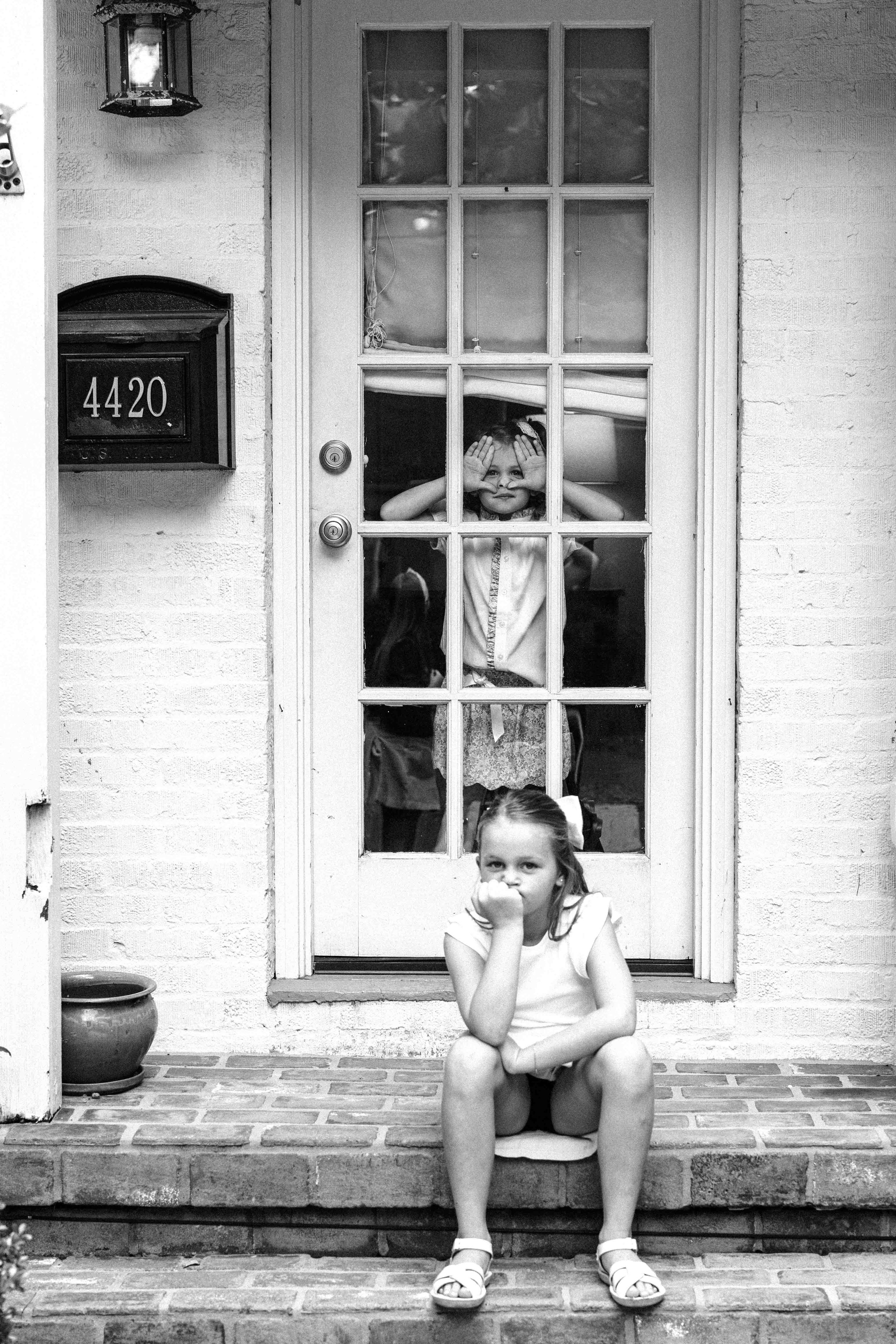 A girl sitting on the steps outside a house, with two other children behind a glass-paned door looking out. One child is peeking through the window with hands framing her face, the other child is partially visible behind her. The girl in the foregrou