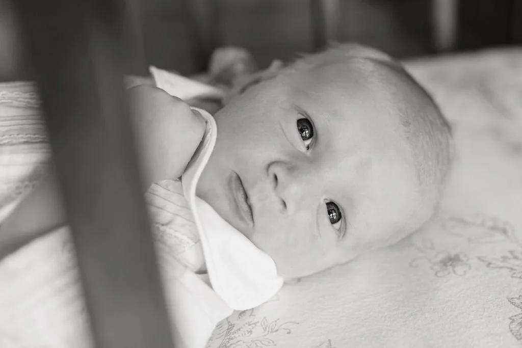 A black and white photo of a baby lying on a bed, looking at the camera, with a neutral expression.