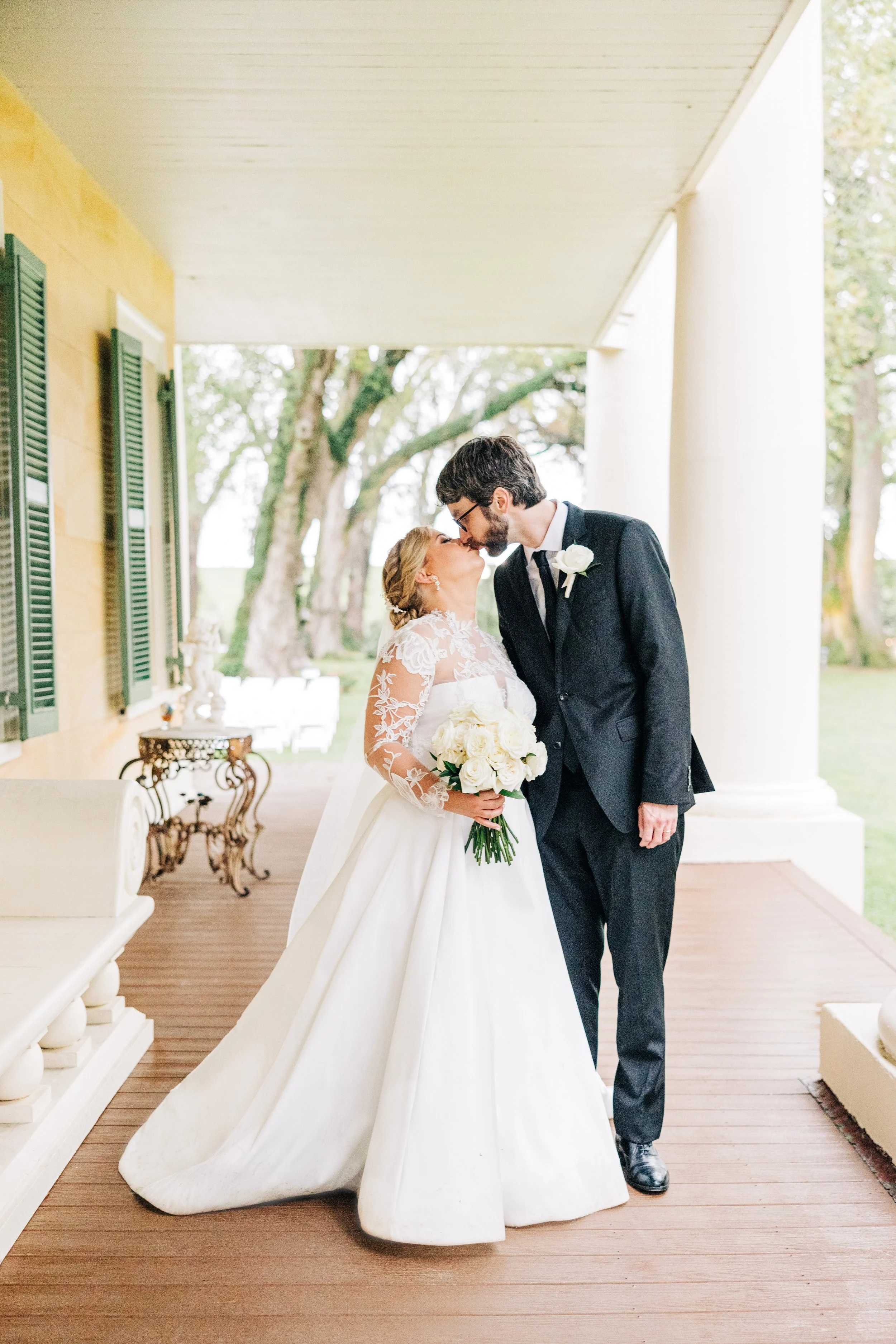 A newlywed couple sharing a kiss on the porch of a house, with the bride holding a bouquet of white roses, and the groom dressed in a black suit with a white flower boutonniere.