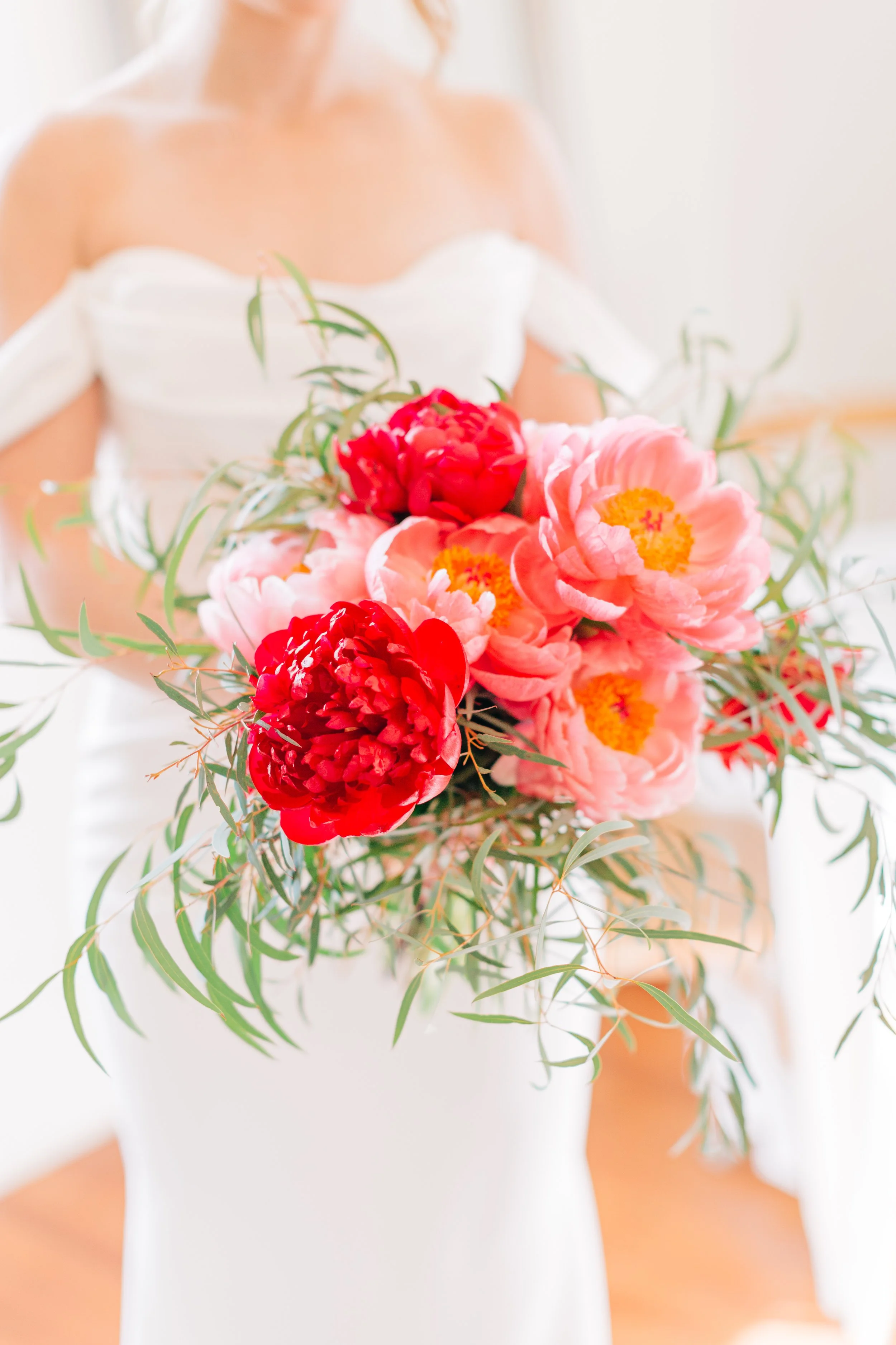 A woman in a white dress holding a bouquet of pink and red peonies and greenery.