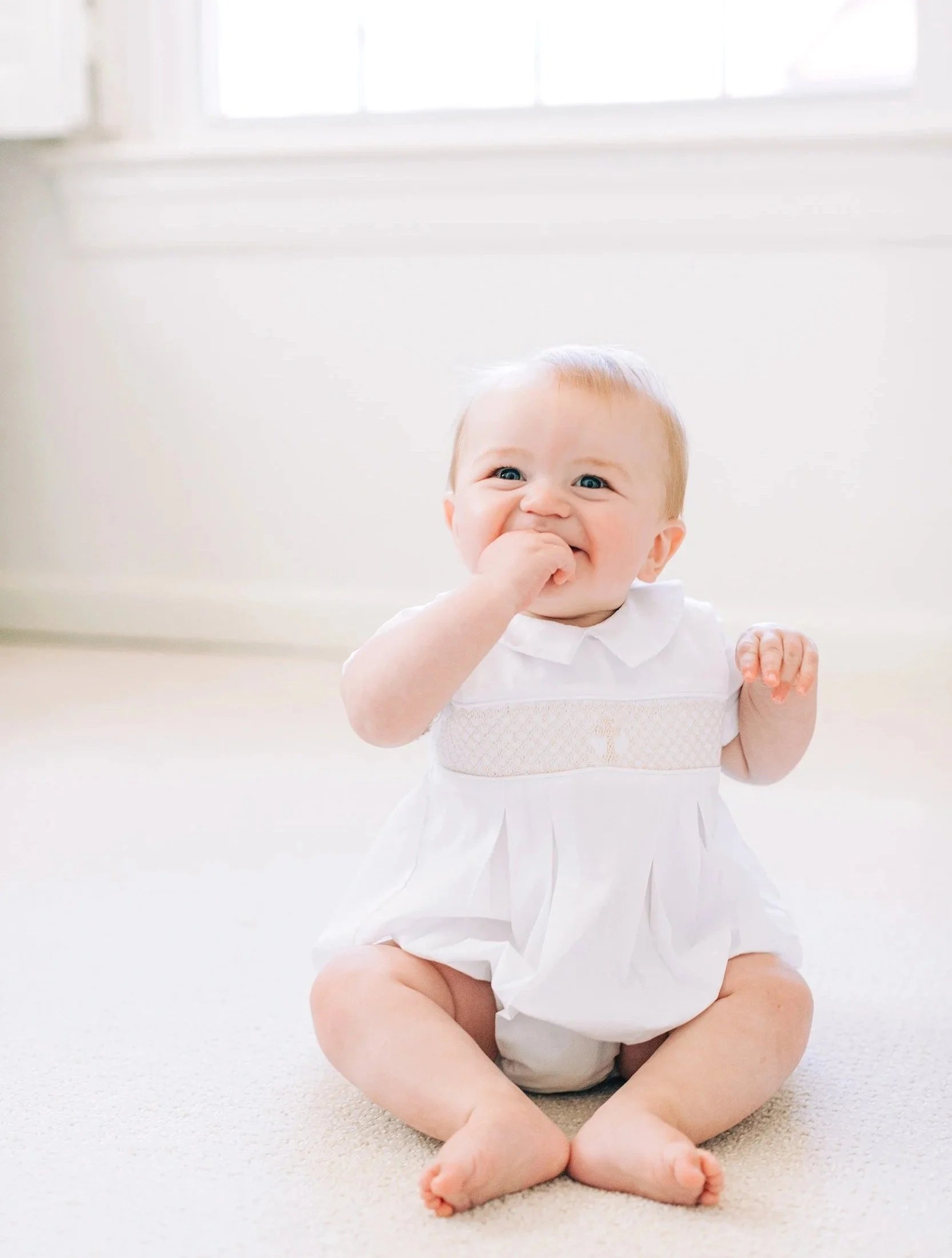 A baby sitting on a cream-colored carpet, wearing a white dress, with hand in mouth and smiling.