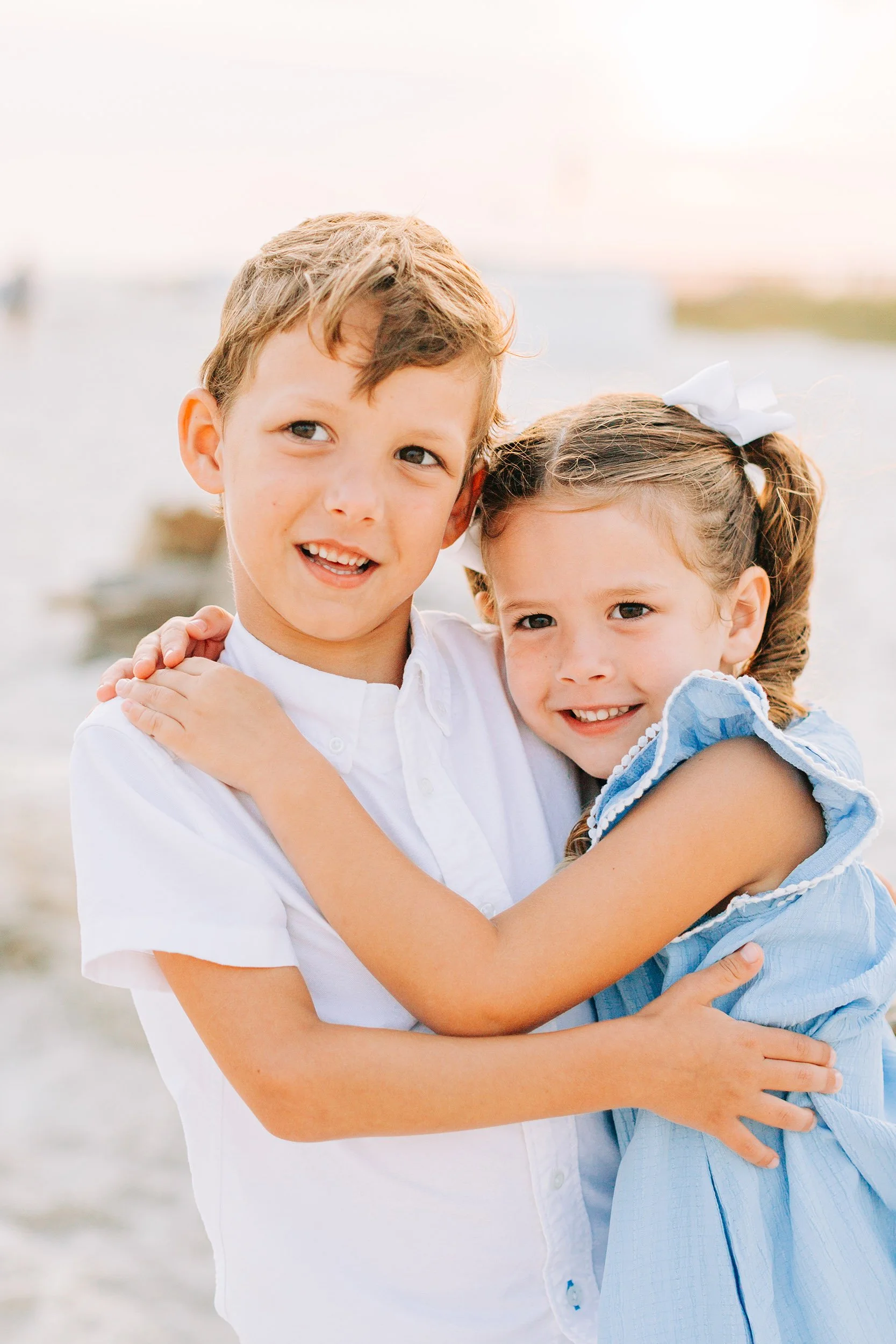 Two smiling children, a boy and a girl, hugging at the beach during sunset.