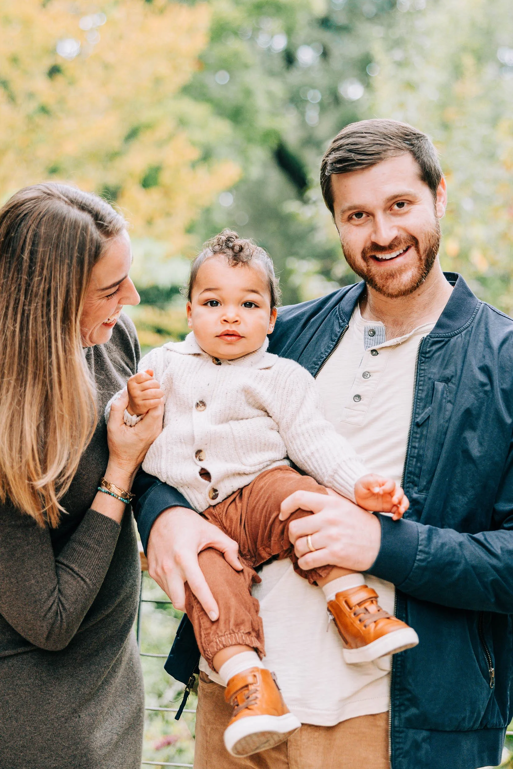 A happy family enjoying outdoor time, consisting of a woman, a man, and a young child, with blurred trees in the background.