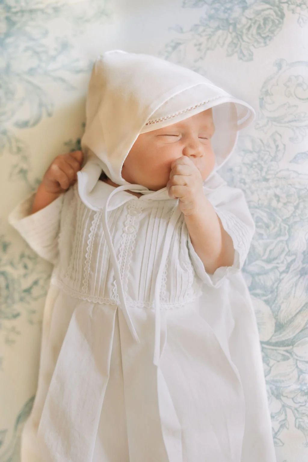 A newborn baby sleeping on a pillow, dressed in a white outfit with a matching white bonnet, clutching their hand near their face.