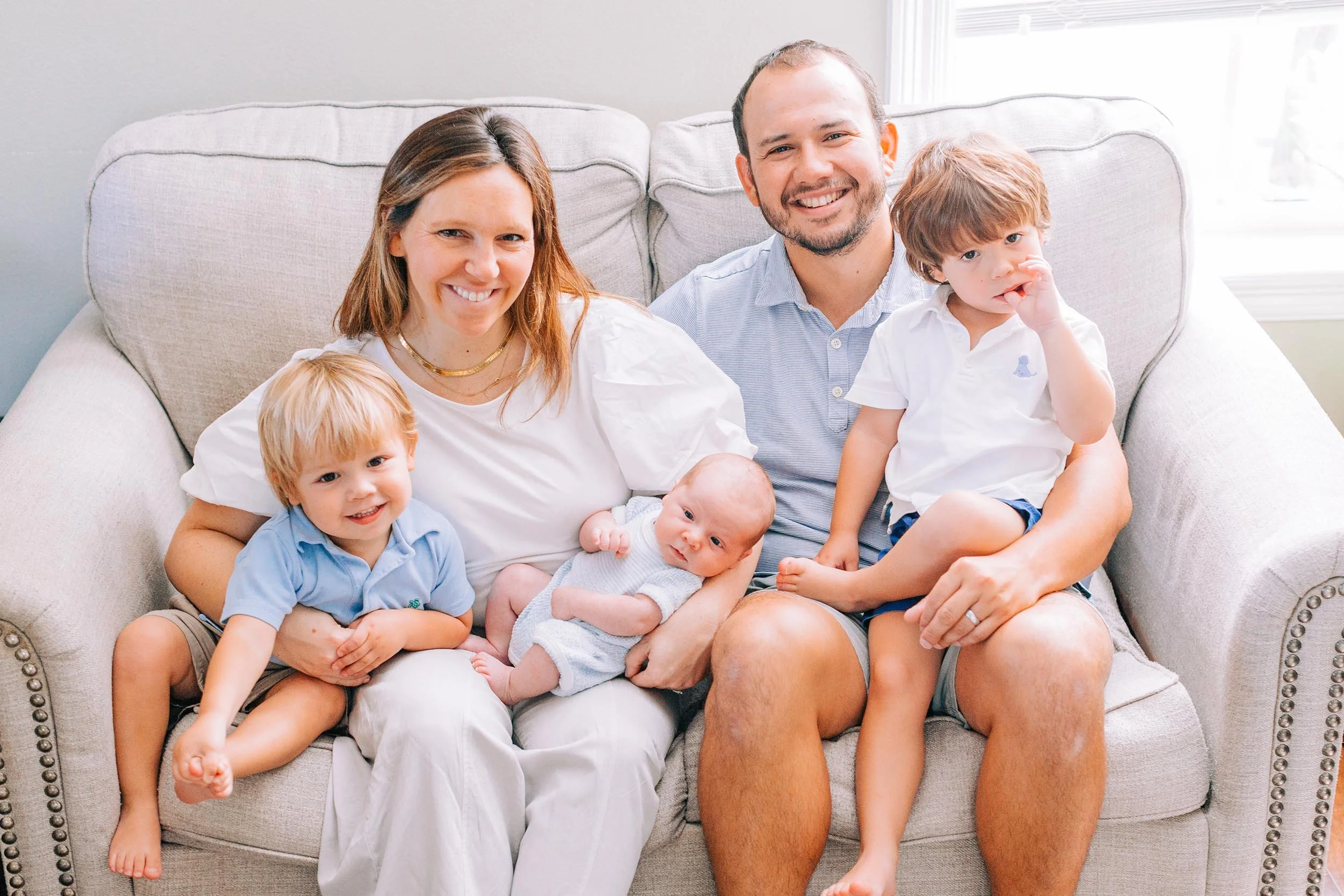 A family sitting on a sofa in a bright living room. The family includes a woman, a man, and three young children. The children are sitting on the adults' laps or close by, and everyone is smiling or looking at the camera.