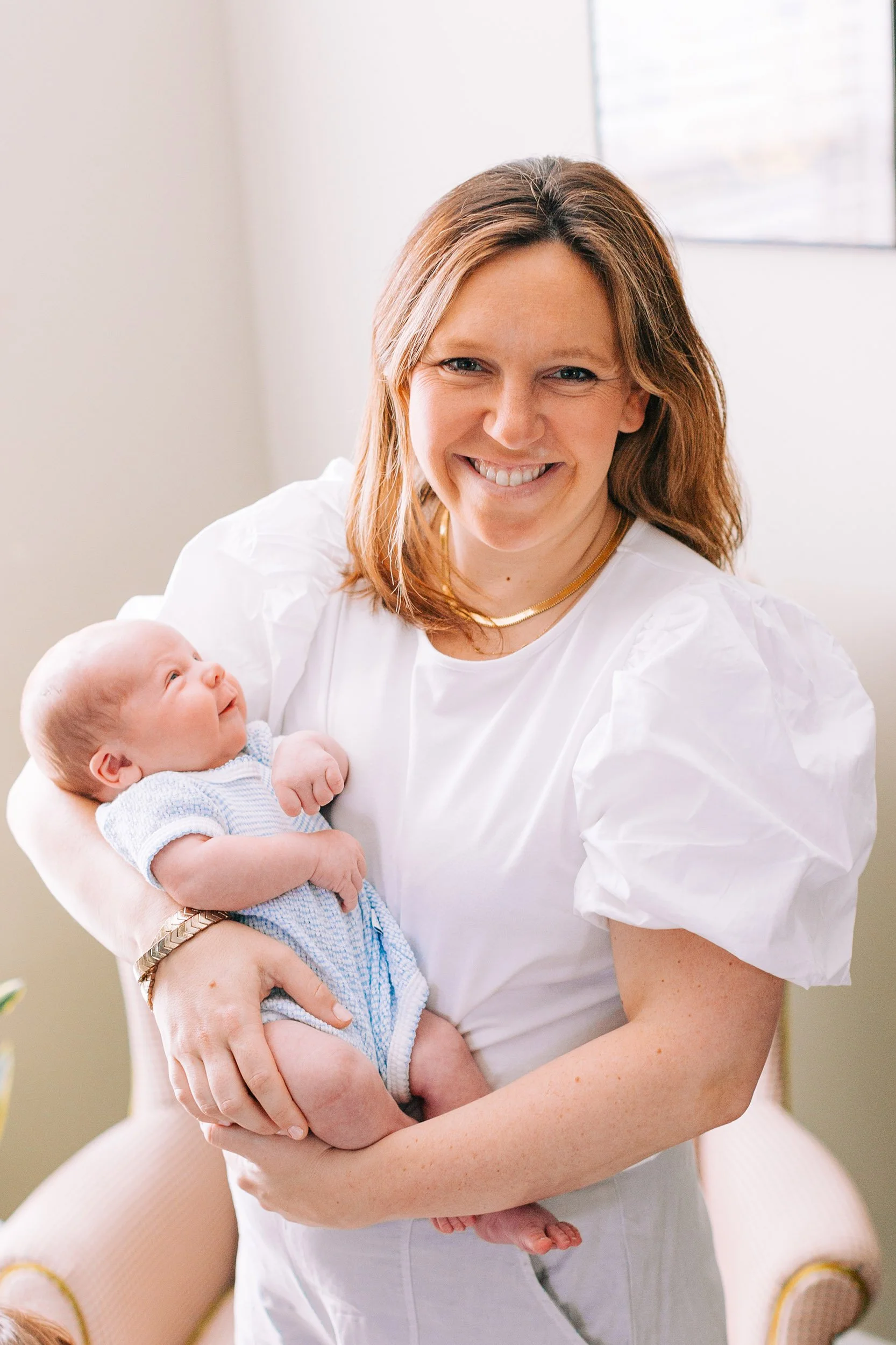 A woman with shoulder-length hair and a gold necklace holding a smiling newborn baby dressed in a blue and white outfit, in a brightly lit room.