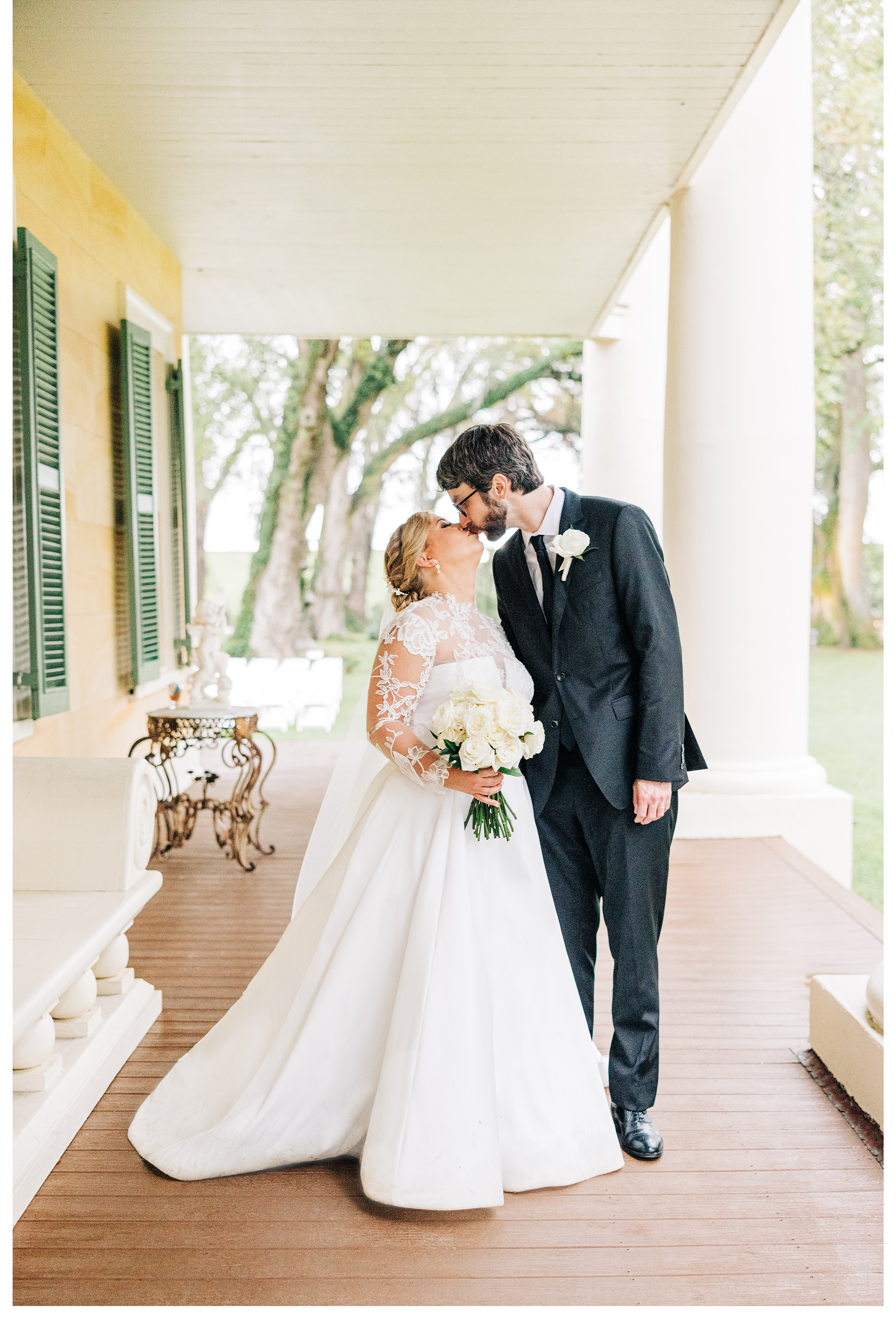 A bride and groom are sharing a kiss on a porch with white columns and green trees in the background. The bride is holding a bouquet of white roses and wearing a white wedding dress with long lace sleeves. The groom is dressed in a dark suit with a white shirt and a black tie.