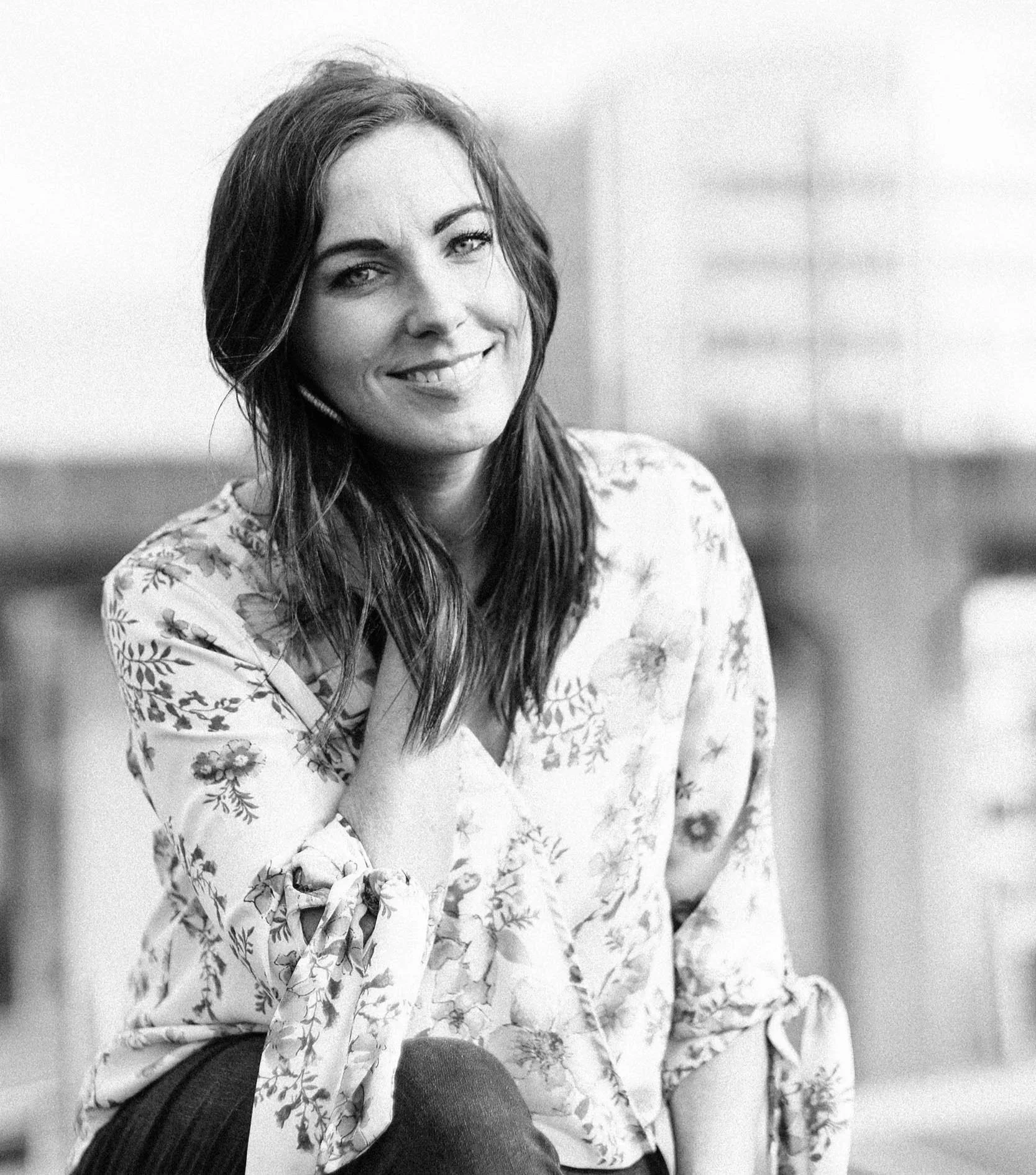 A woman with long hair smiling, sitting indoors wearing a floral patterned blouse and dark tights.