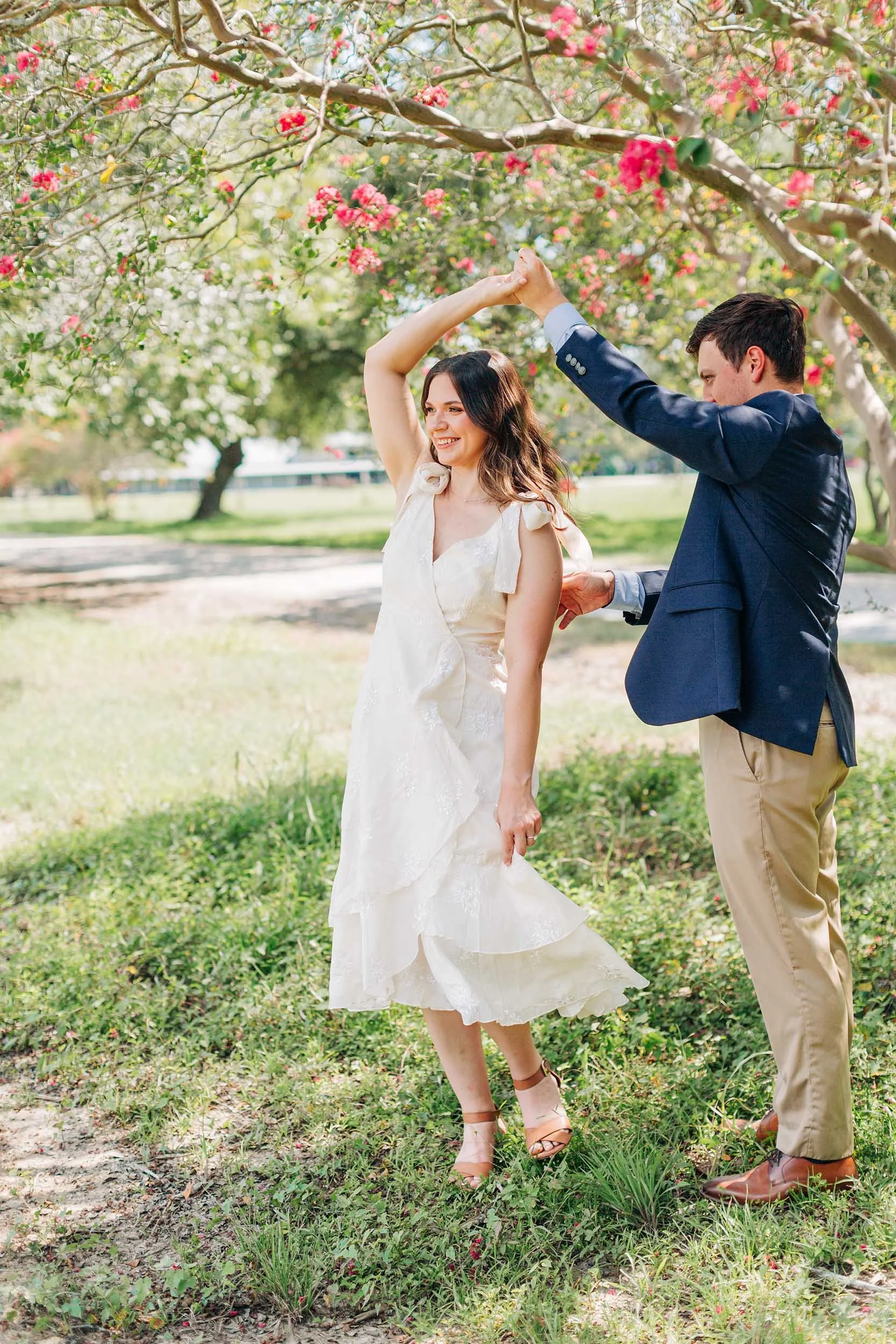 A couple dancing outdoors in a park with pink blossoming trees, the woman wearing a white dress and the man in a suit jacket and khakis.