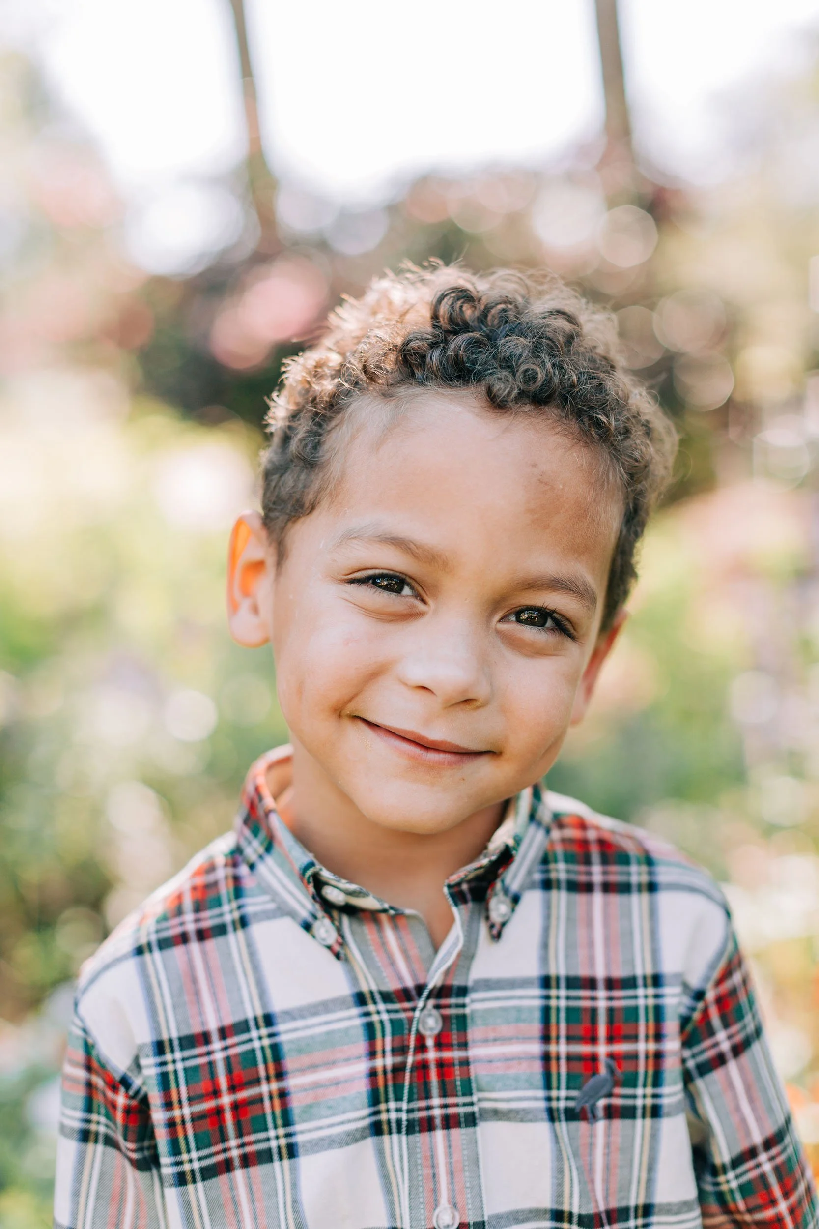 A young boy with curly brown hair and a bright smile, wearing a plaid shirt, standing outdoors with blurred trees and sunlight in the background.