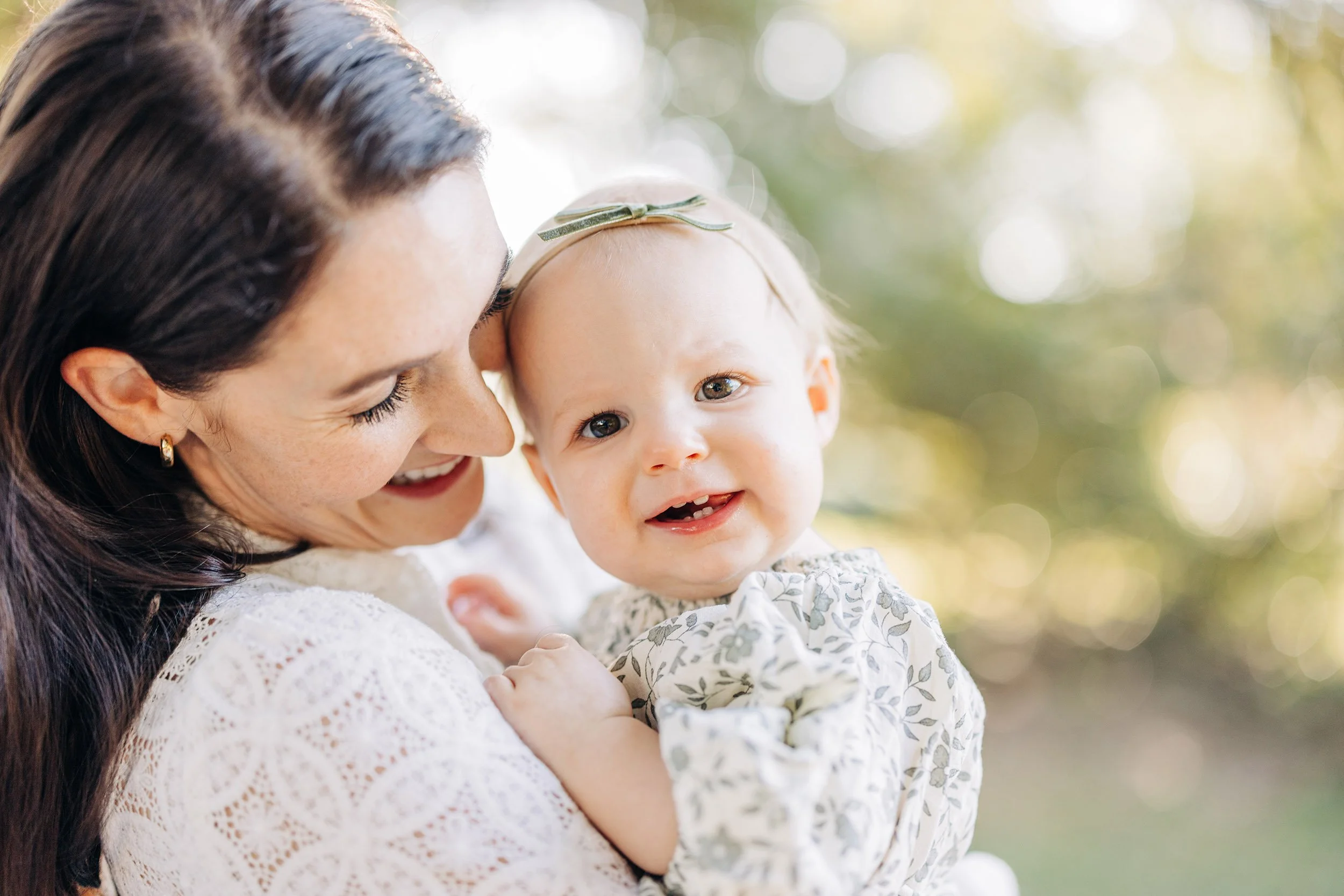 A woman with long dark hair and earrings holds a baby girl with a bow headband, both smiling outdoors with blurred sunlight and trees in the background.