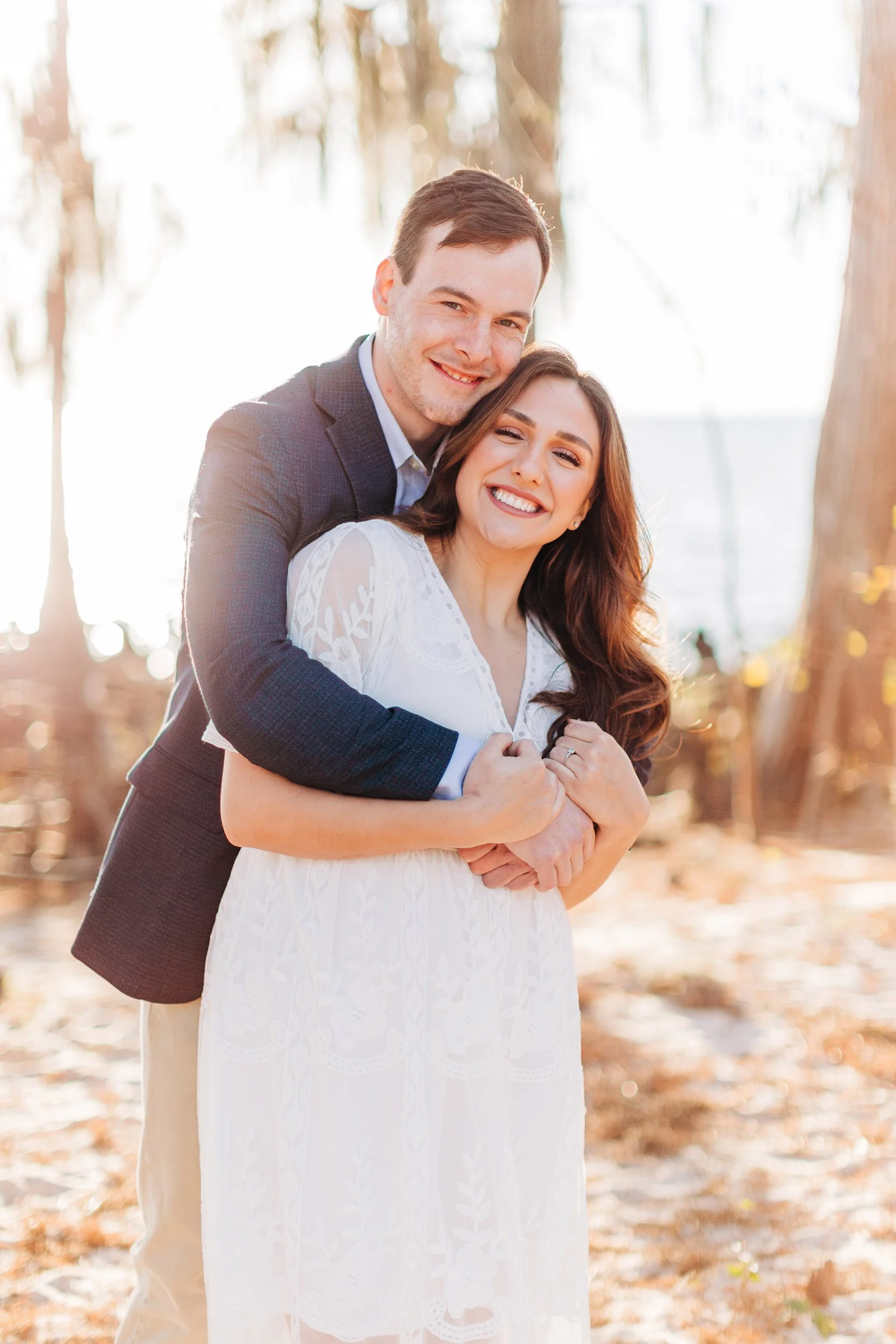 A smiling couple embracing outdoors with autumn leaves and trees in the background, sunlight shining through.
