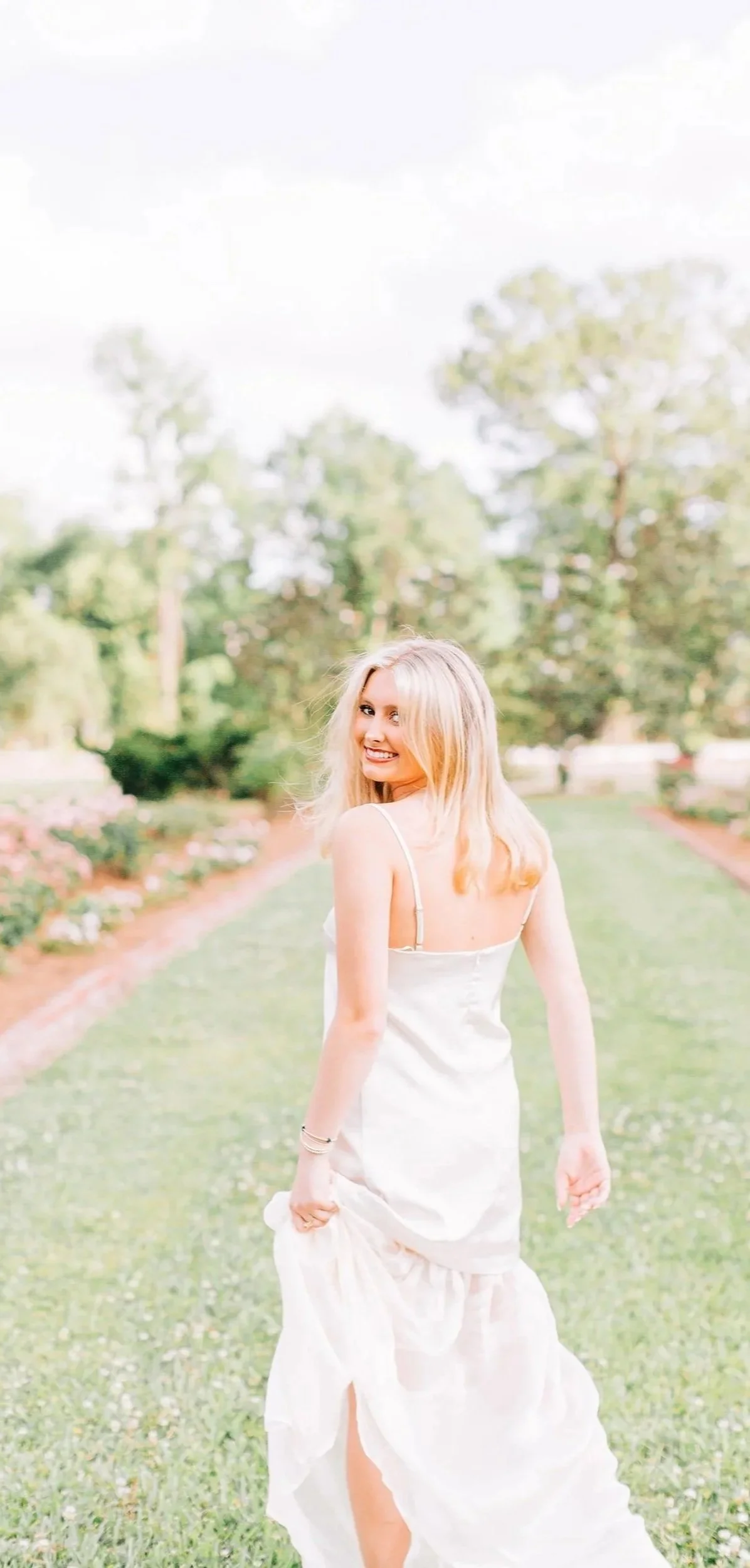 A young woman with blonde hair wearing a light-colored dress, smiling and walking outdoors on a grassy path with trees and flowers in the background.