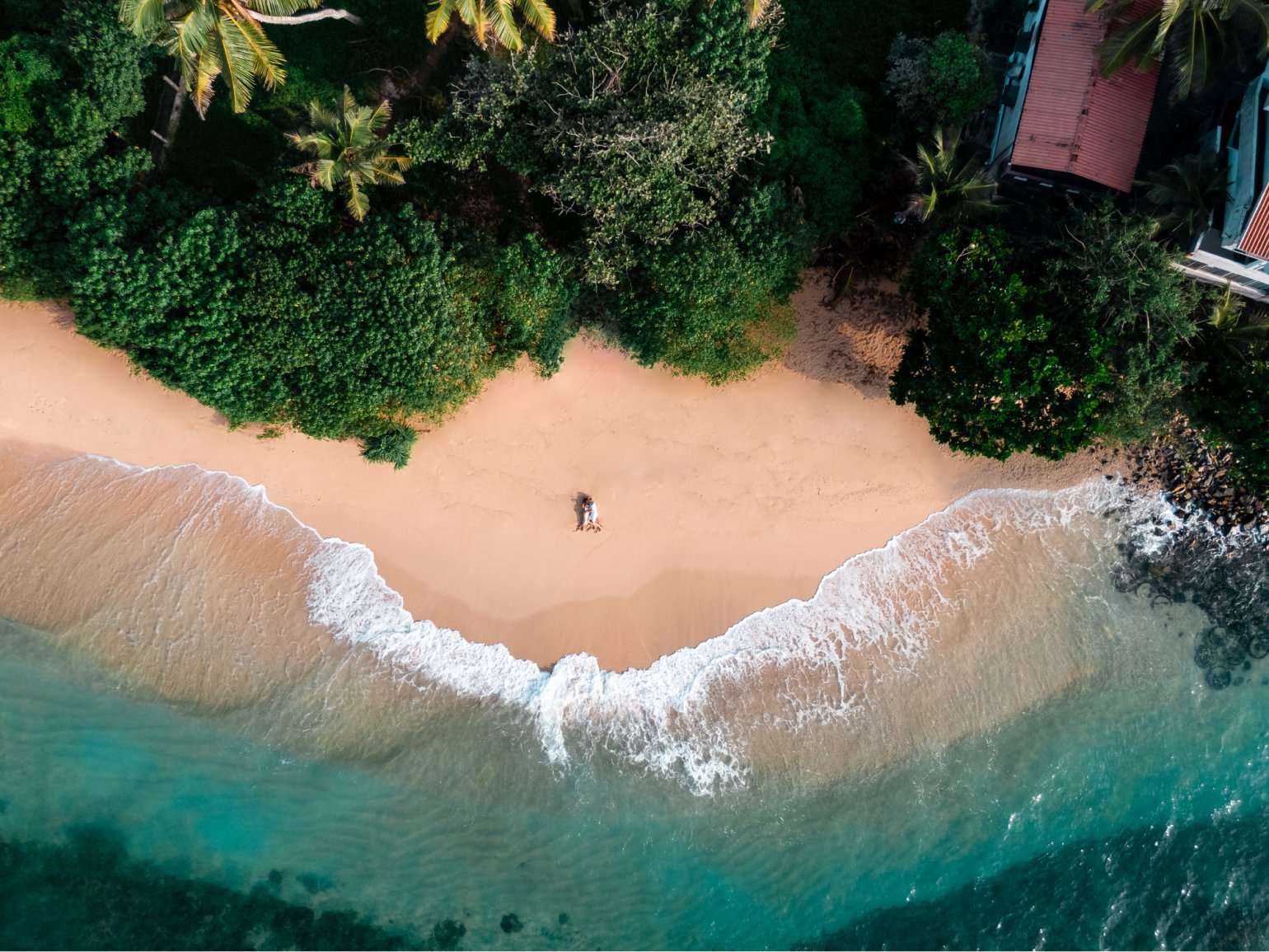 Aerial photograph of a newlywed couple laying down in the sand of an empty beach, surrounded by palmtrees and other vegetation.