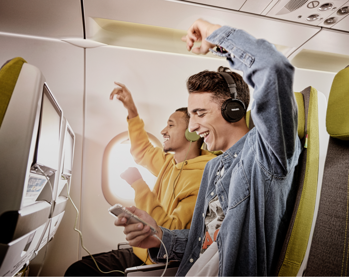Two smiling young men sit in the Economy Class seats of a TAP airplane, listening to music and celebrating with their arms in the air.