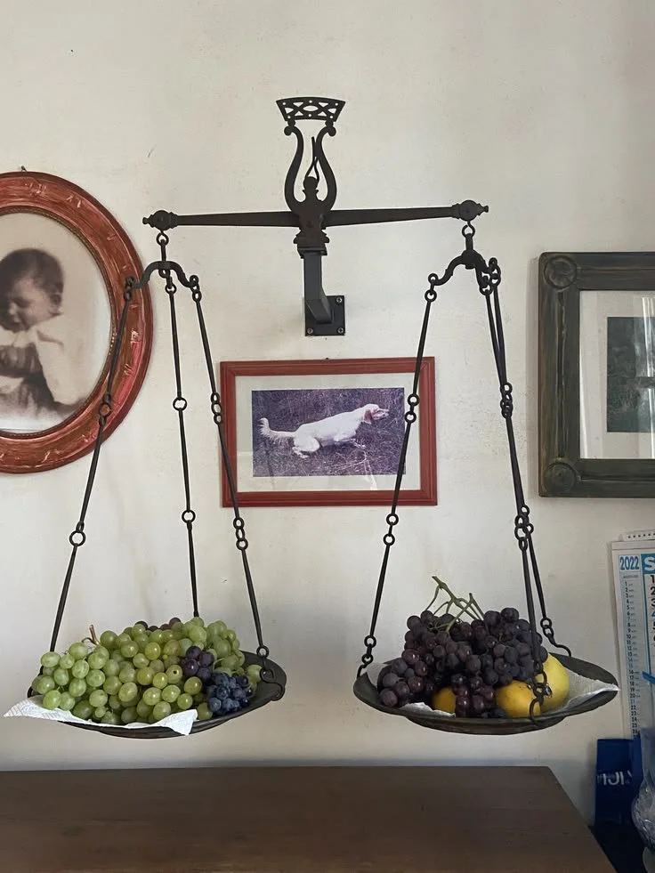 A hanging scale with two pans, one holding green grapes and the other holding dark grapes and lemons, mounted on a wall with framed pictures.