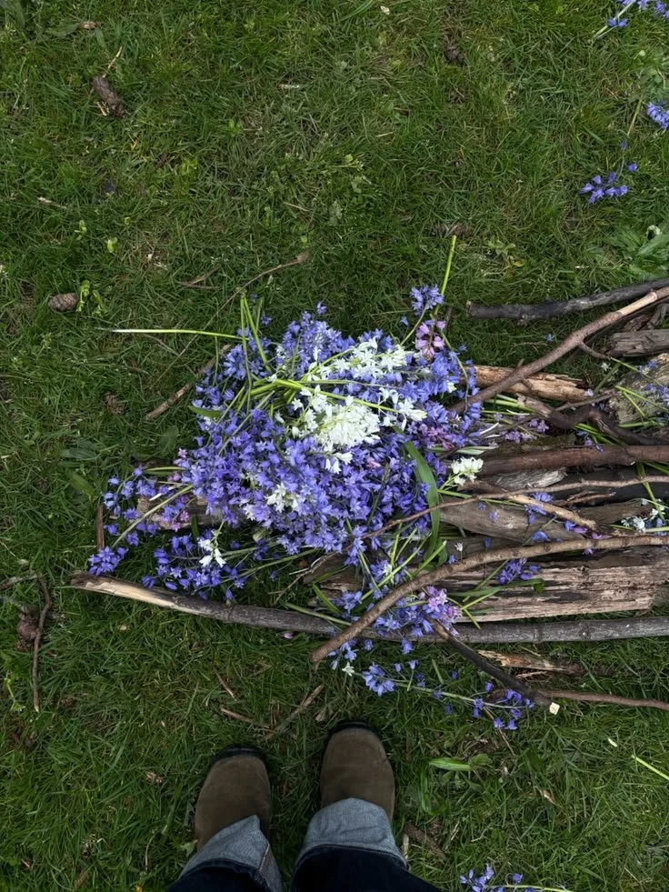 A bundle of purple and white flowers, possibly lavender or similar, lying on green grass with some sticks and twigs around, and the person's shoes visible at the bottom of the image.