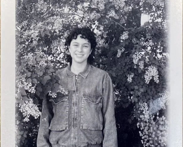 A young woman with short curly hair smiling and wearing a denim jacket standing in front of flowering bushes.