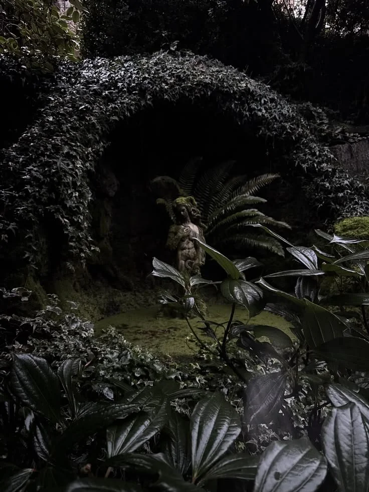 A dark, lush garden scene with dense greenery and foliage. At the center, there is a sculpture of a classical male figure surrounded by ferns and moss-covered plants, partially hidden by dark leaves.