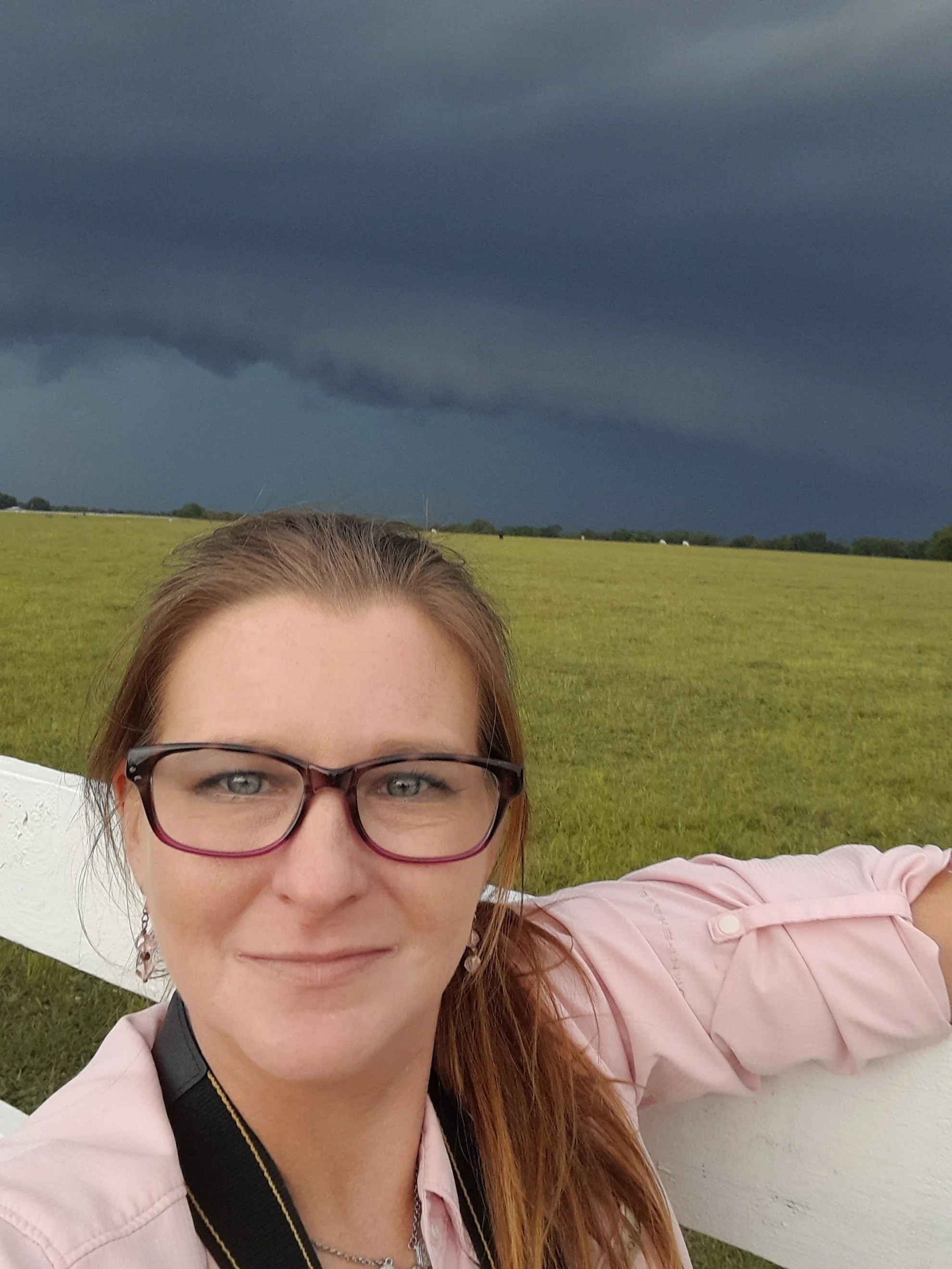A woman with red hair, glasses, and a pink shirt, taking a selfie in front of a field with a dark storm cloud in the sky.