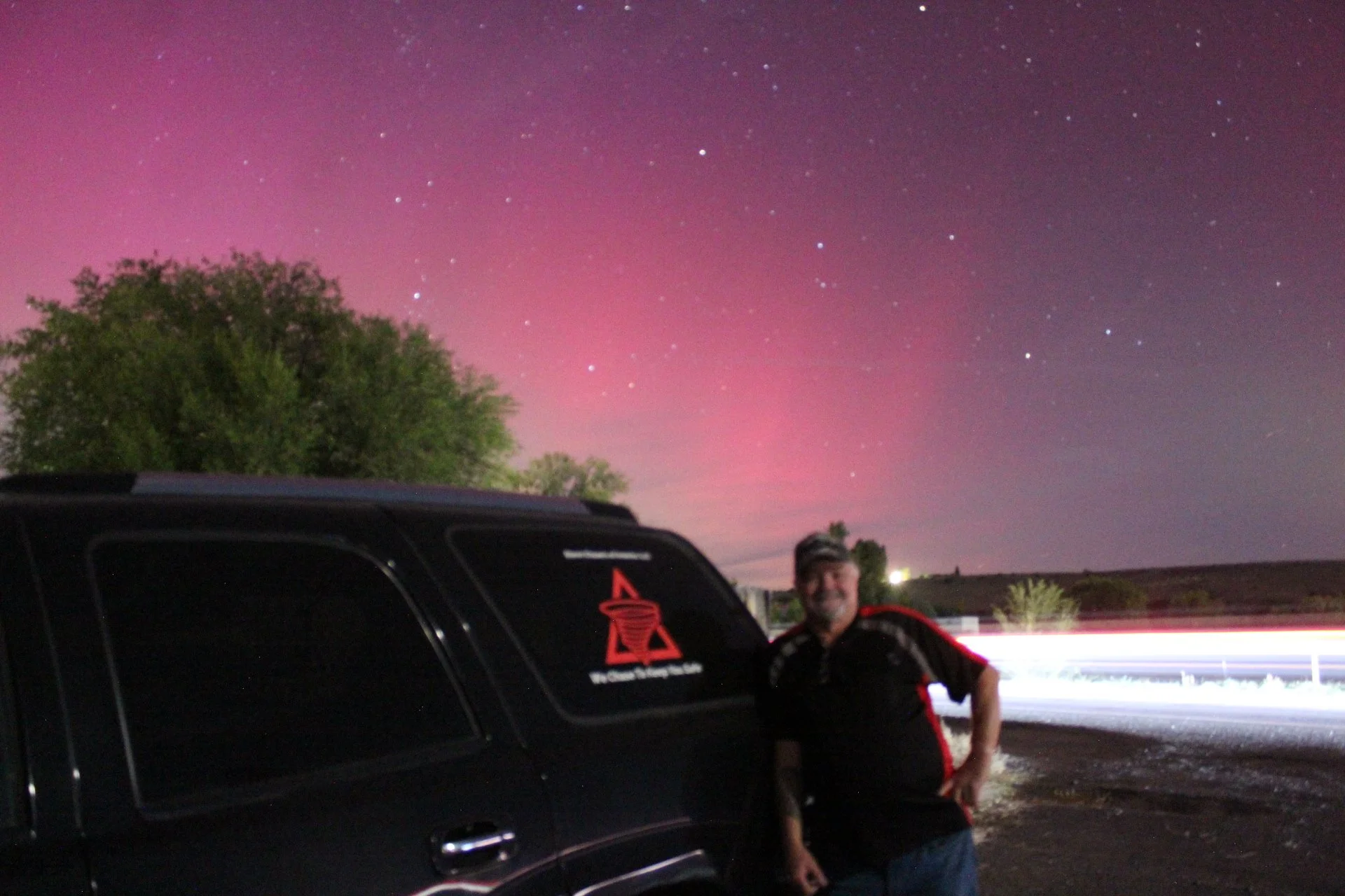 Night sky with pink aurora and stars, person standing next to parked SUV.