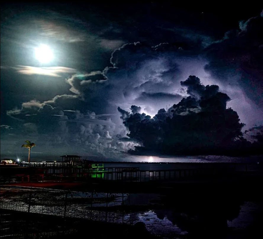 Night scene of a dramatic sky over a waterfront, featuring the moon illuminating clouds, a palm tree, and a silhouette of a dock. Lightning illuminates the clouds, creating a striking contrast between the bright moon and stormy scenery.