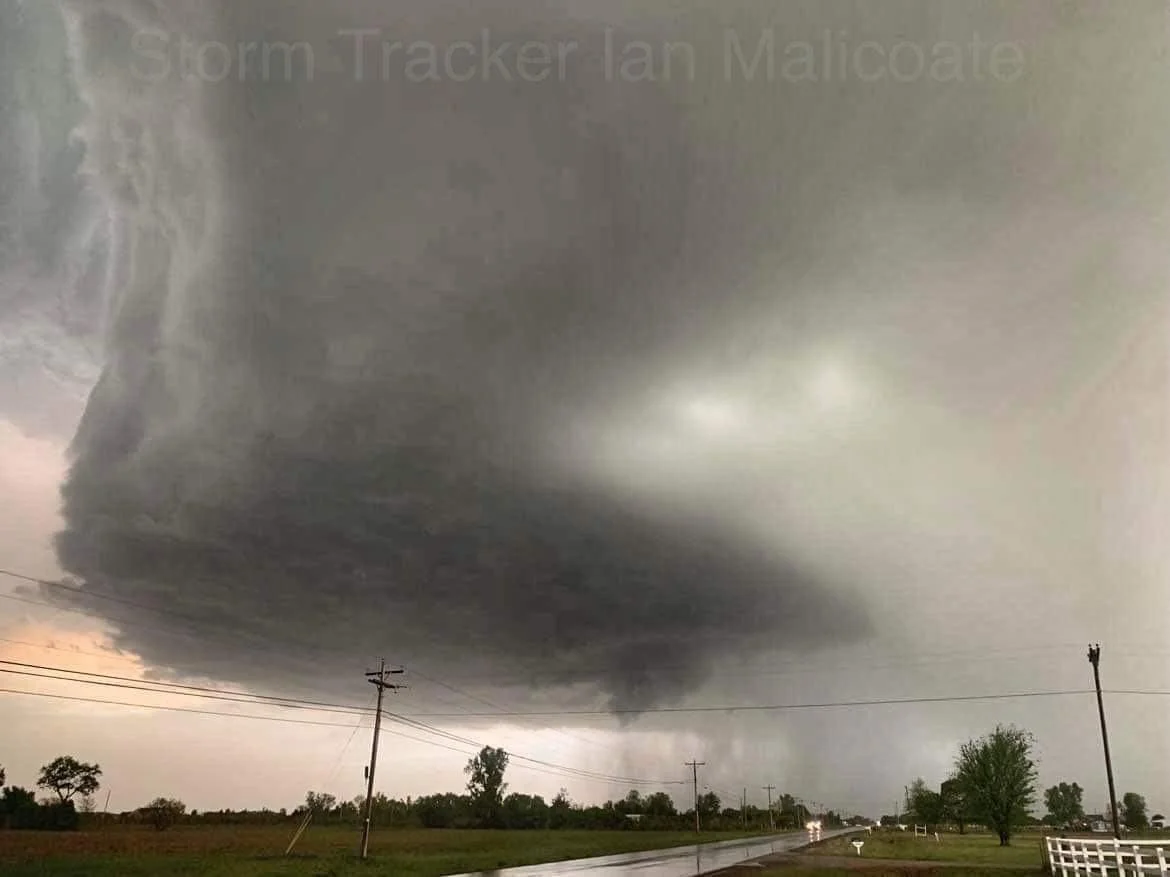 Large storm cloud forming over a rural road, with dark skies and surrounding fields, indicating severe weather conditions.