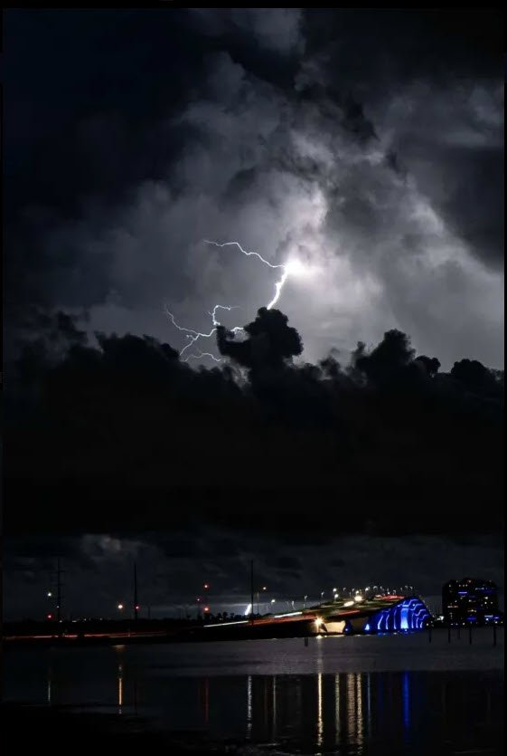 Lightning storm over a city bridge at night with illuminated clouds and bright lights reflecting on water below.