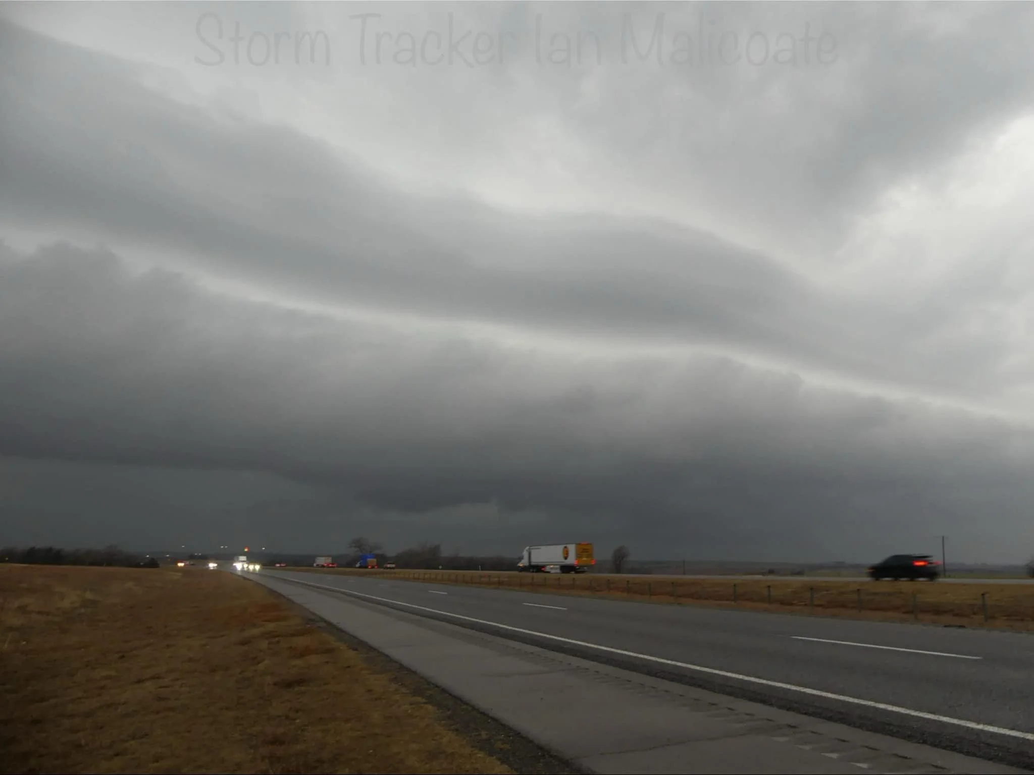 Overcast sky with dark storm clouds above a highway. Vehicles, including a truck and cars, are visible on the road. The landscape is flat and grassy, indicating an open rural area.