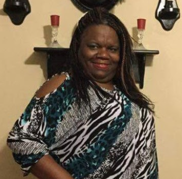 Smiling woman wearing a patterned blouse standing indoors in front of a shelf with decorative items and candle holders.