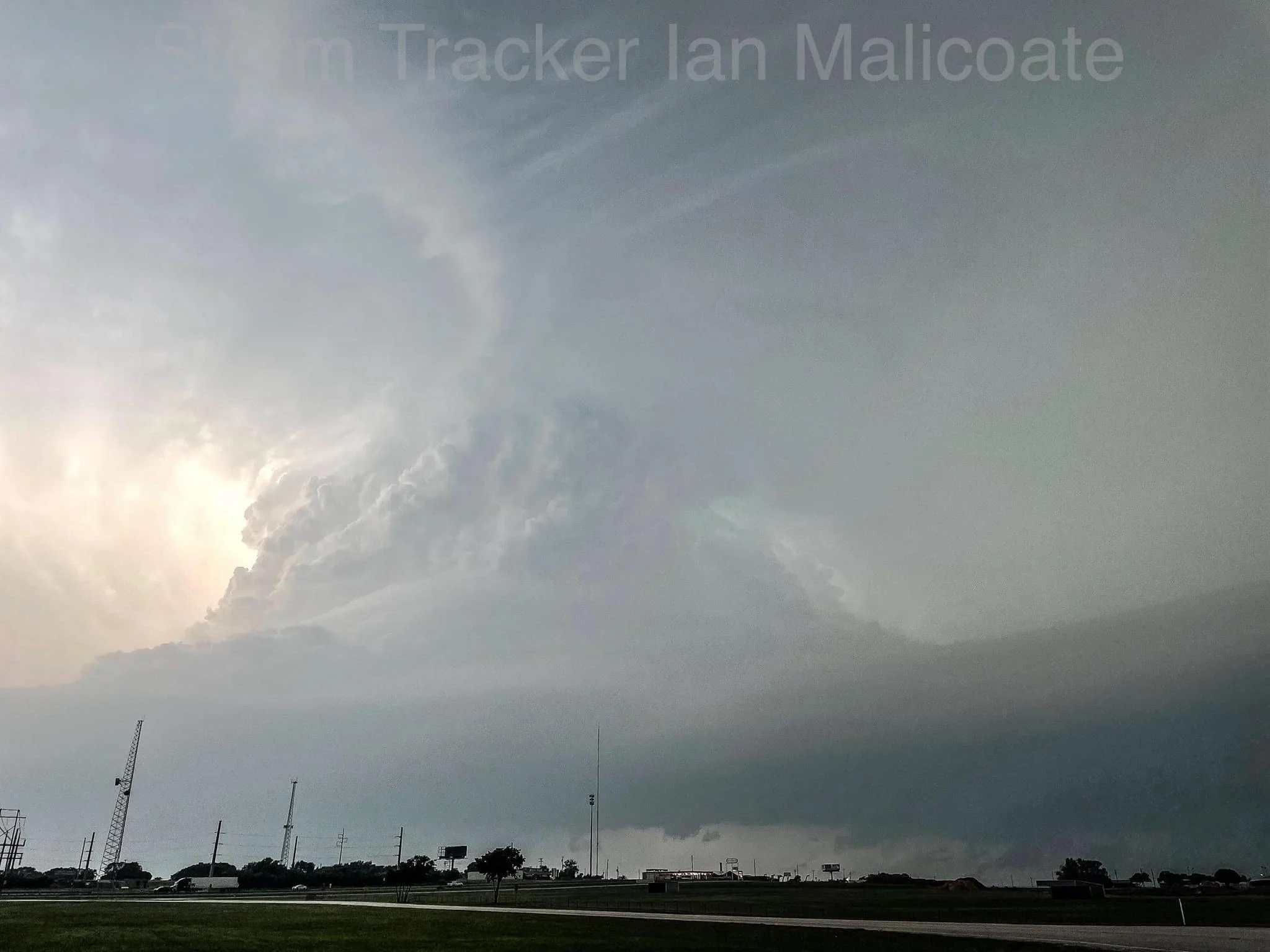 Massive storm cloud formation with dark skies and some sun peeking through, over a flat landscape with visible transmission towers and trees.