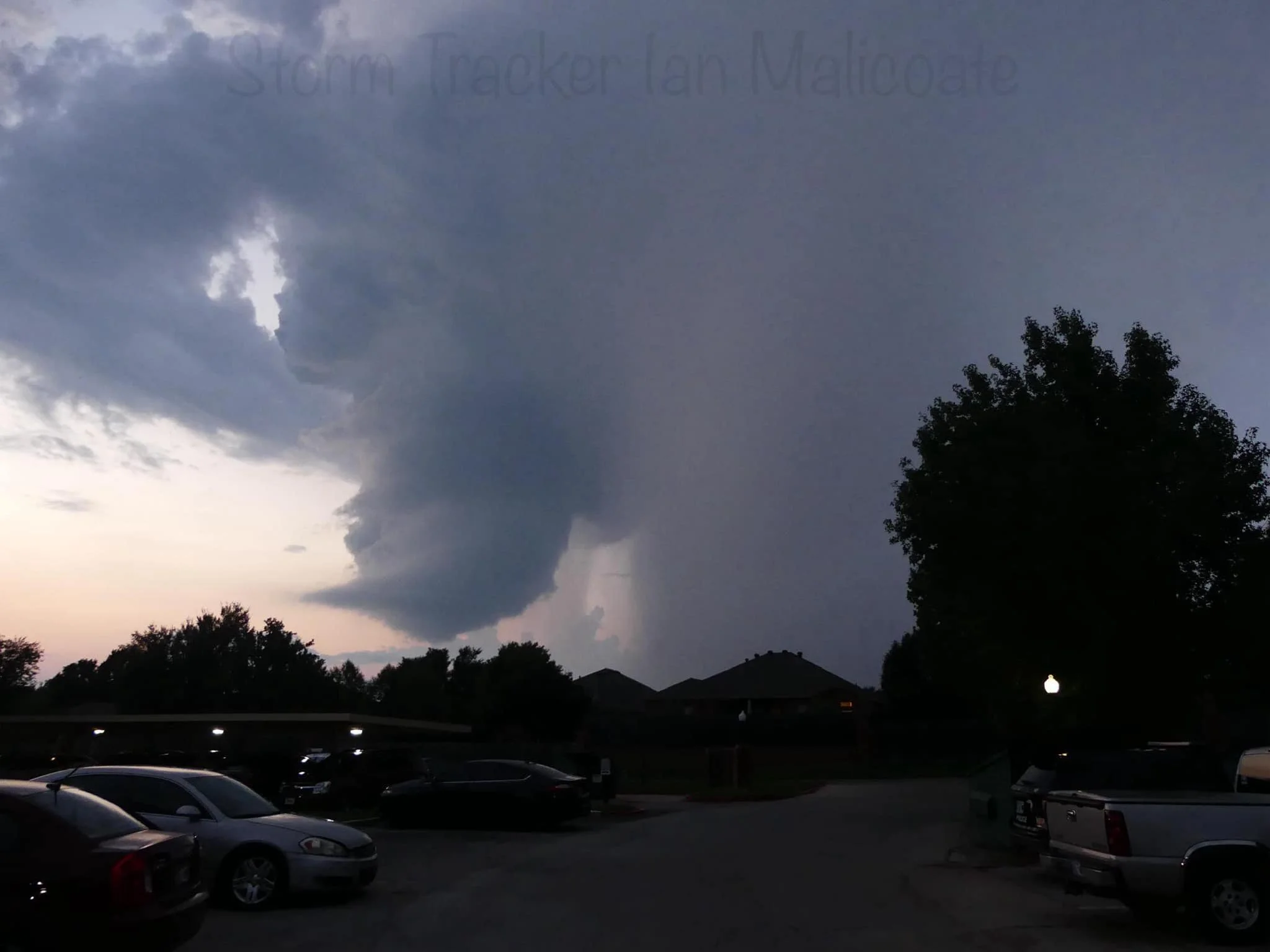 Dark storm cloud forming in the evening sky above a parked row of cars and trees, with a faint sunset in the background.