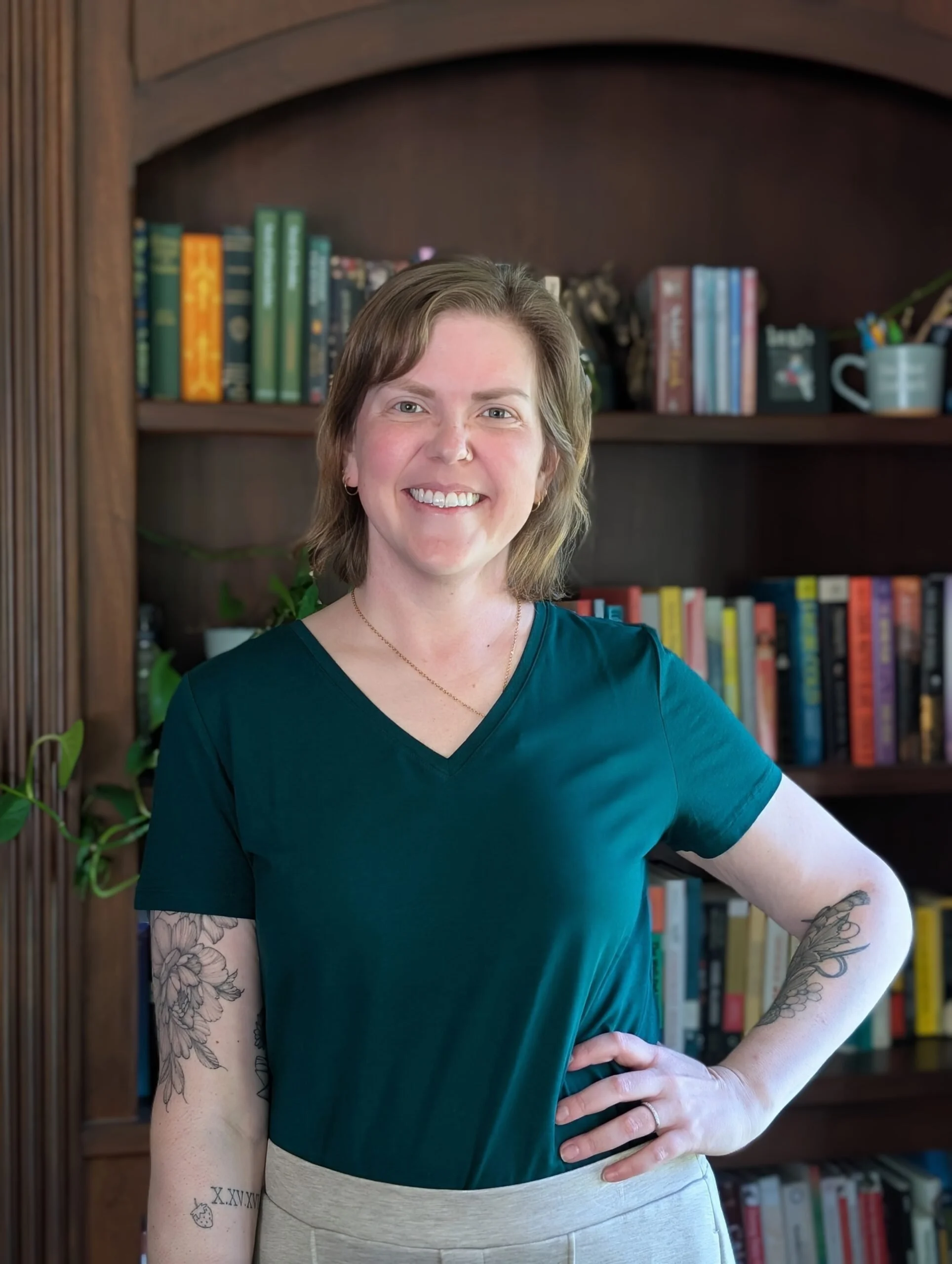 A woman with short hair, tattoos on her forearms, and a necklace, standing in front of a bookshelf filled with books and a mug holding pens, smiling at the camera.