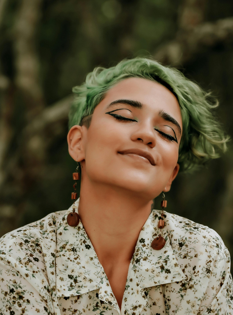 A woman with short, curly green hair smiling with eyes closed, wearing floral shirt and long earrings, outdoors with blurred trees in the background.
