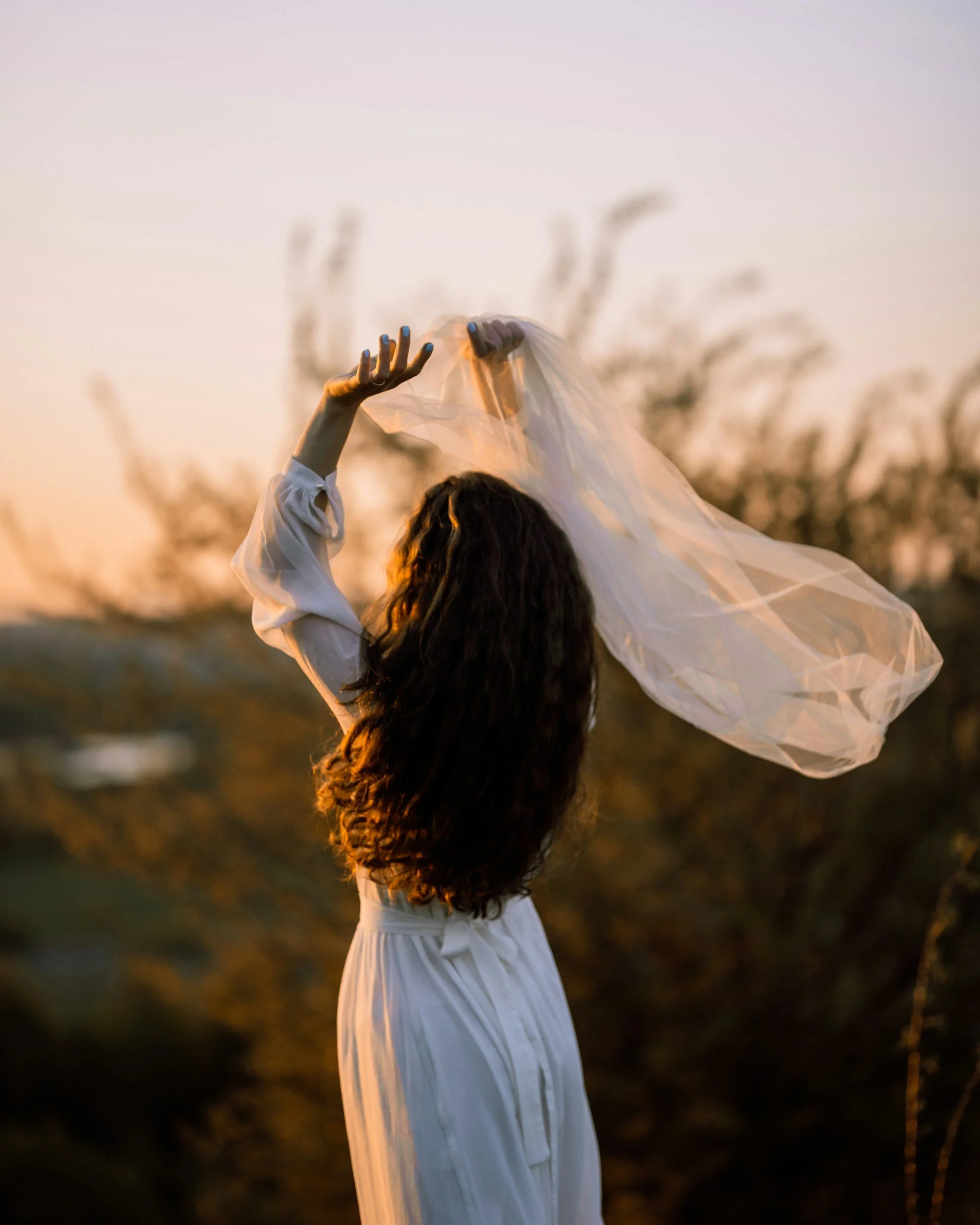A woman with long, curly hair wearing a white dress holding a sheer cloth up in a field during sunset. Trauma-informed therapist in Portland Oregon providing individual and couples therapy
