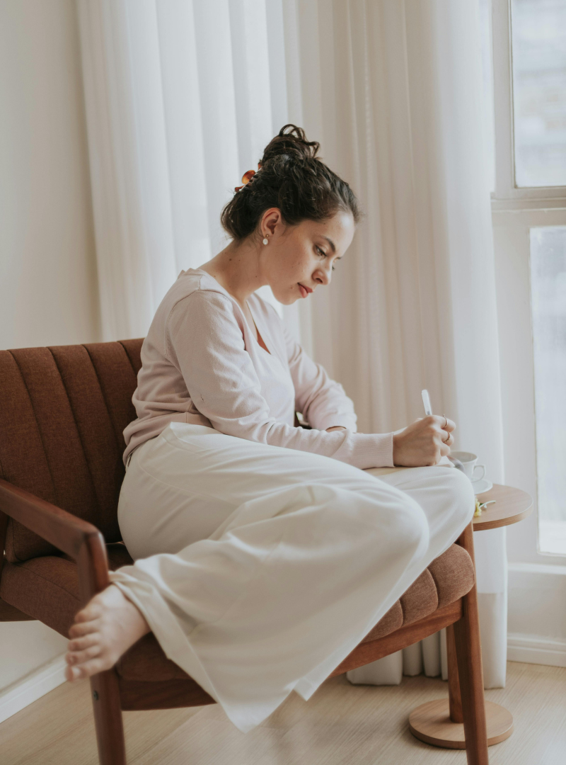 A woman with dark hair in a bun, wearing a light pink sweater and white pants, sitting on a brown sofa by a window, writing in a notebook. Trauma-informed therapist in Portland Oregon providing individual therapy