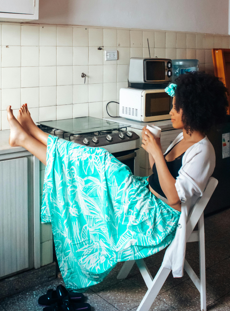 A woman with curly hair, wearing a white cardigan, black top, and a turquoise patterned skirt, sits on a chair in a kitchen with her legs resting on the counter, drinking from a mug.