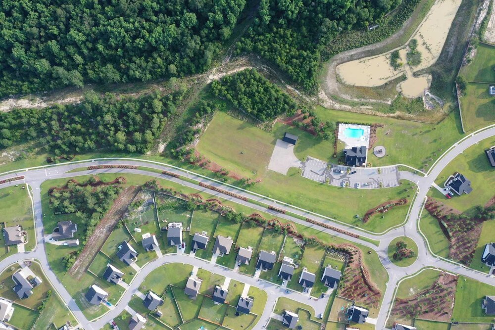 Aerial view of a master-planned residential community showcasing a well-manicured streetscape with detailed perimeter landscaping, a central roundabout, and a large amenity center with a pool, parking lot and club house.