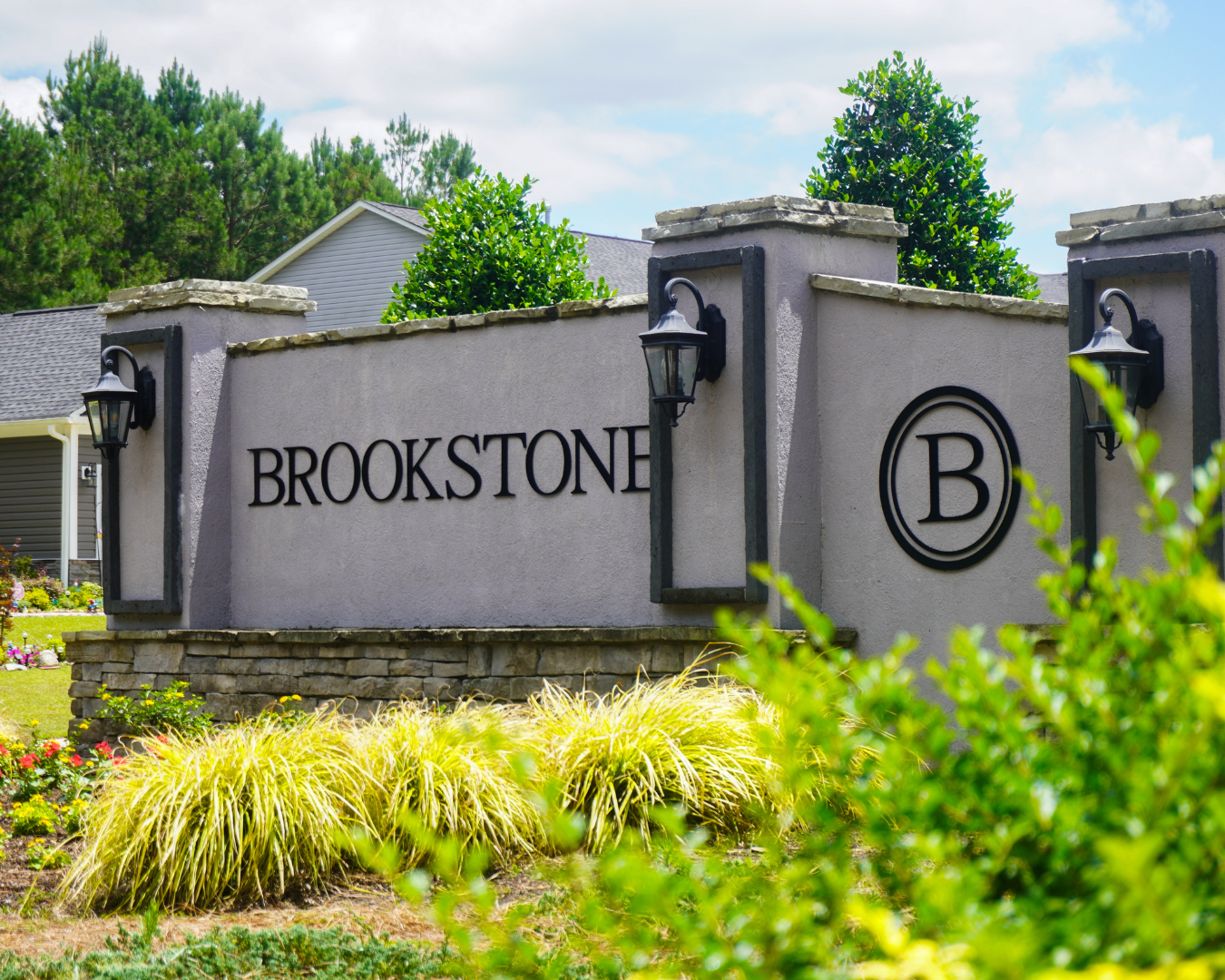 Brick and stucco entrance monument for Brookstone neighborhood featuring modern black lettering and decorative lighting, surrounded by manicured landscaping with ornamental grasses and colorful flower beds.