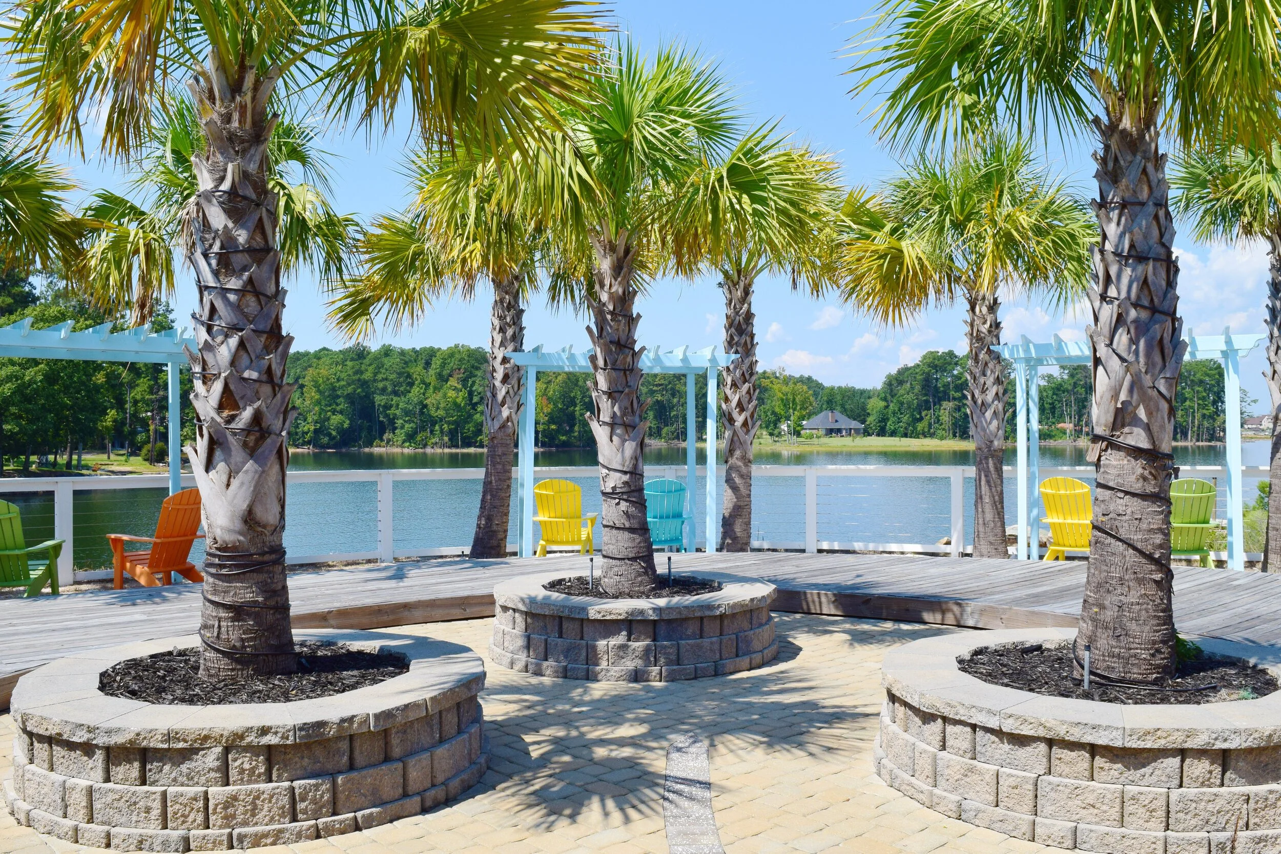 Lakeside patio featuring palm trees planted in raised stone beds, surrounded by paver walkways and pastel blue pergolas. Ideal for showcasing community amenities, waterfront landscaping, or tropical outdoor living spaces in resort-style developments.