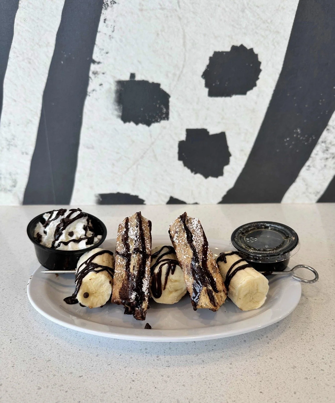 Plate with sliced bananas, cannoli, chocolate-dipped pastries, a small cup of whipped cream with chocolate syrup, and a container of chocolate sauce on a white plate in front of a black and white striped wall.