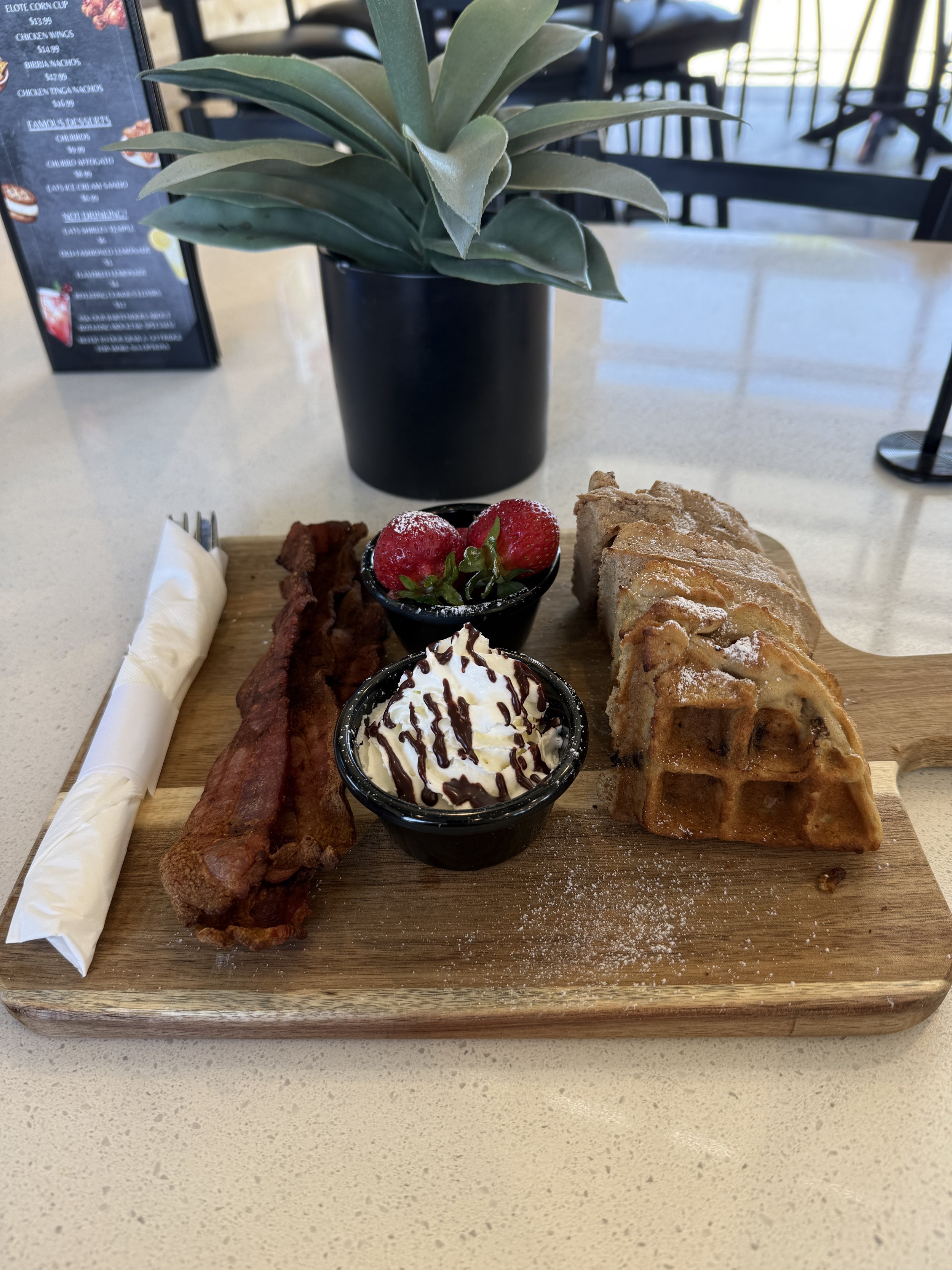 A wooden serving board with a strip of crispy bacon, two strawberries, a slice of apple pie, and a small cup of whipped cream topped with chocolate drizzle. Behind the board is a potted plant with broad green leaves, and there is a black menu stand and some chairs in the background.