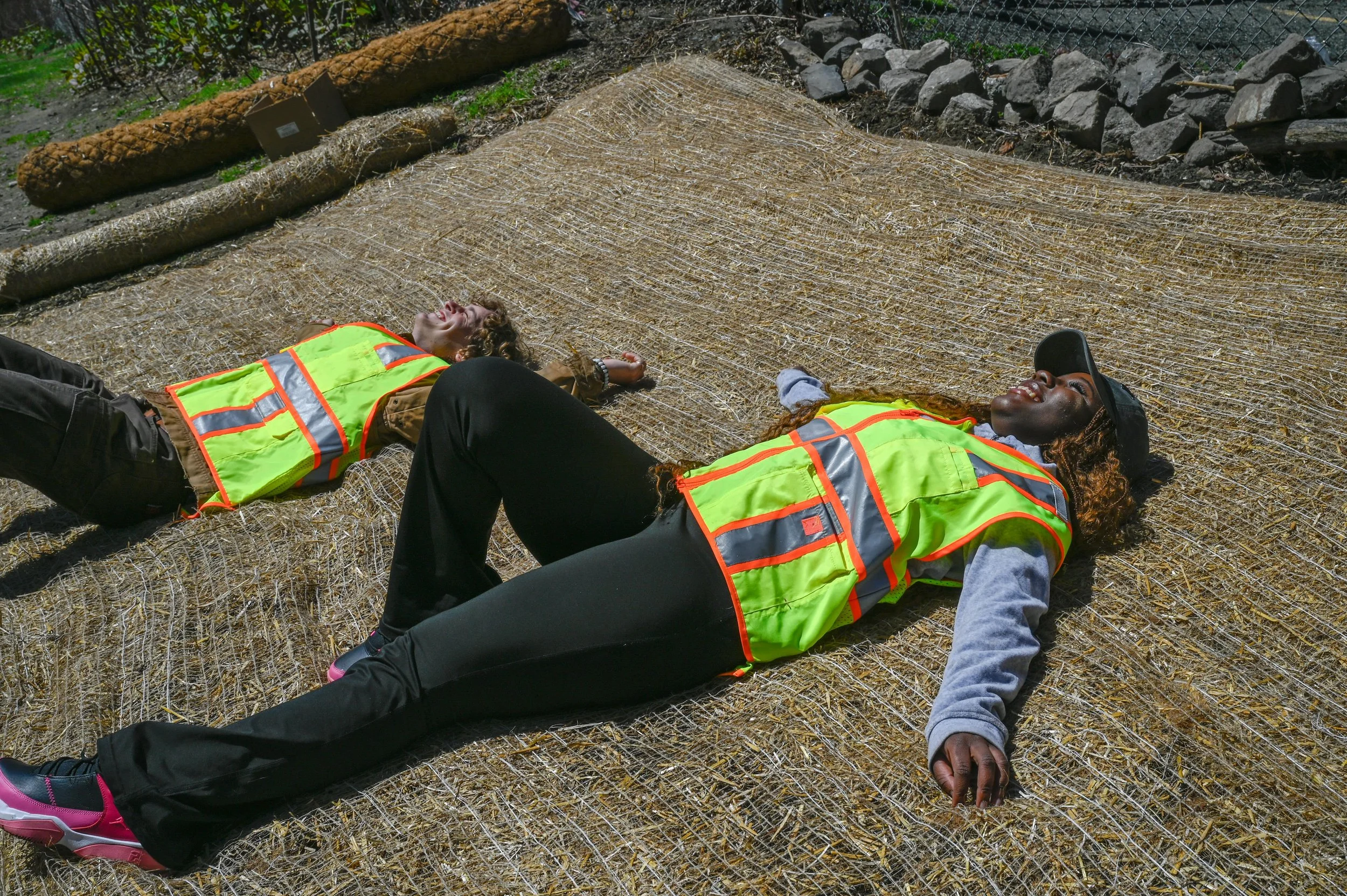 East Boston, MA: April 22, 2024 - Ilaria and Gabriel, two Climate Corps fellows, soak up the sun on an erosion control blanket. A few moments from now they will begin installing straw wattles and plants, just one of the three single-day projects happ