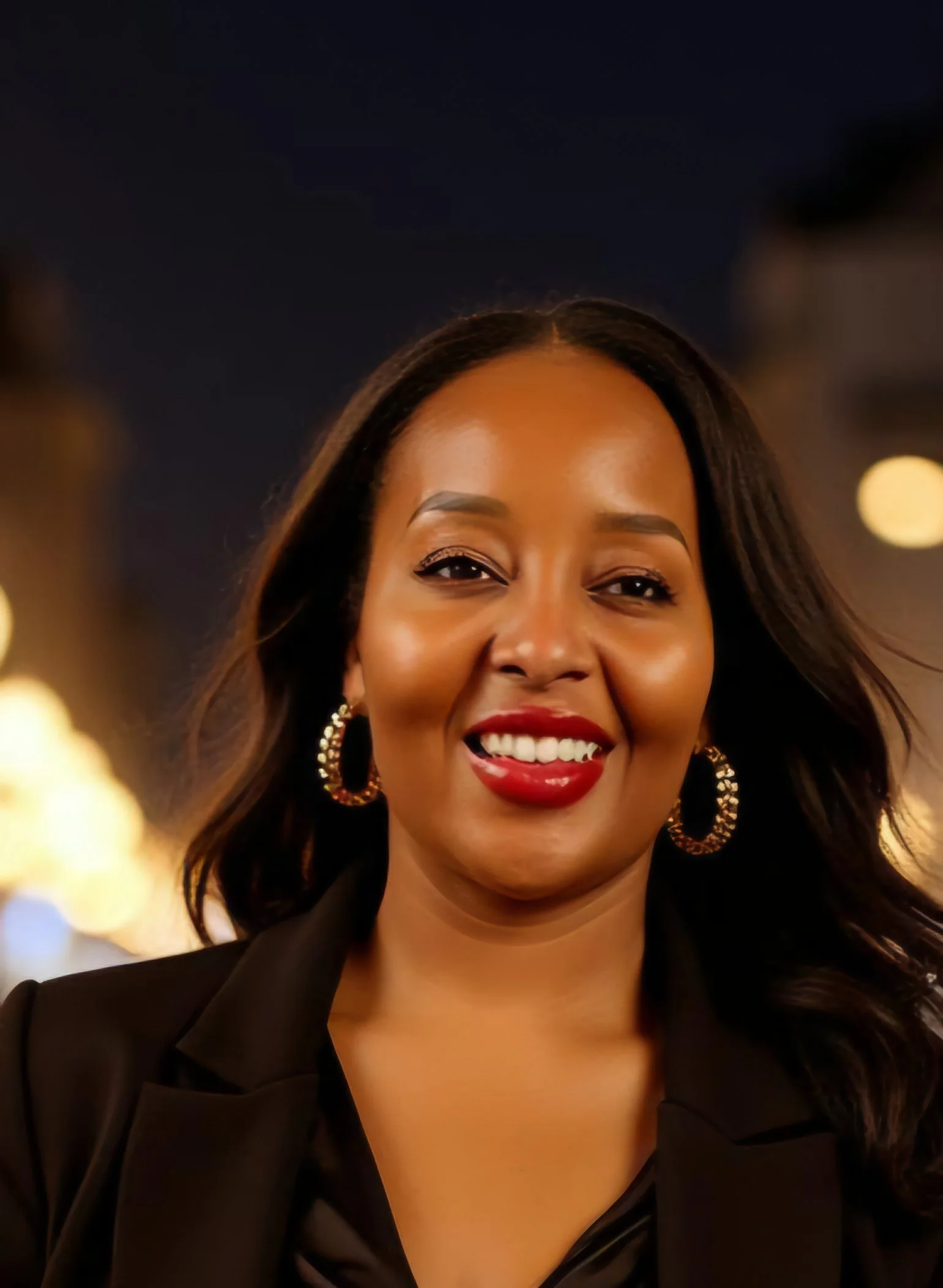 Portrait of a smiling woman with long dark hair, red lipstick, gold hoop earrings, wearing a black blazer, at night with blurred city lights in the background.