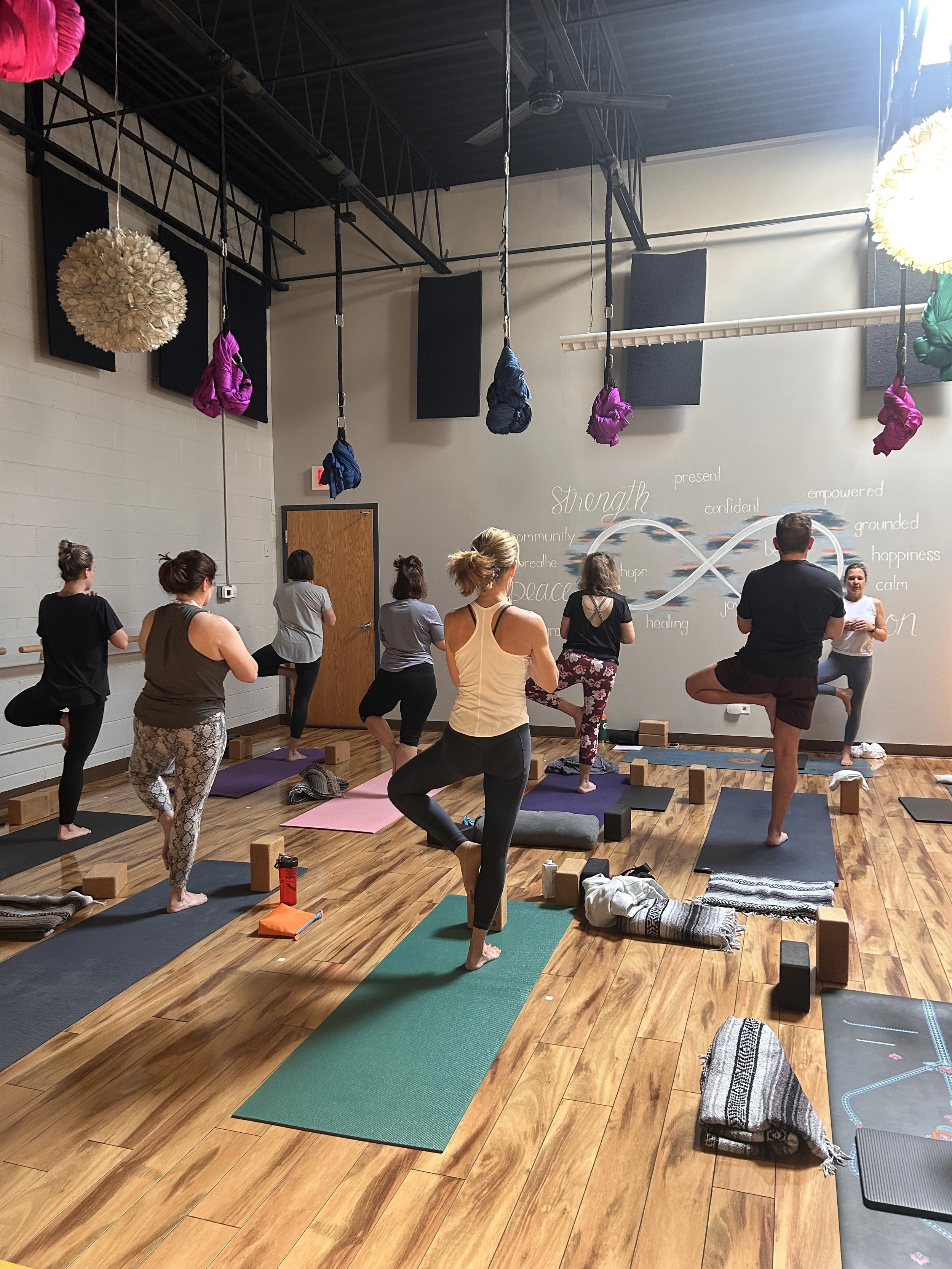 A group of people participating in a yoga or fitness class inside a studio. They are standing on yoga mats, performing balanced poses on one leg. The studio has wooden flooring, black ceiling with hanging purple and turquoise fabric swings, and motivational words like 'strength', 'peace', and 'hope' written on the wall.